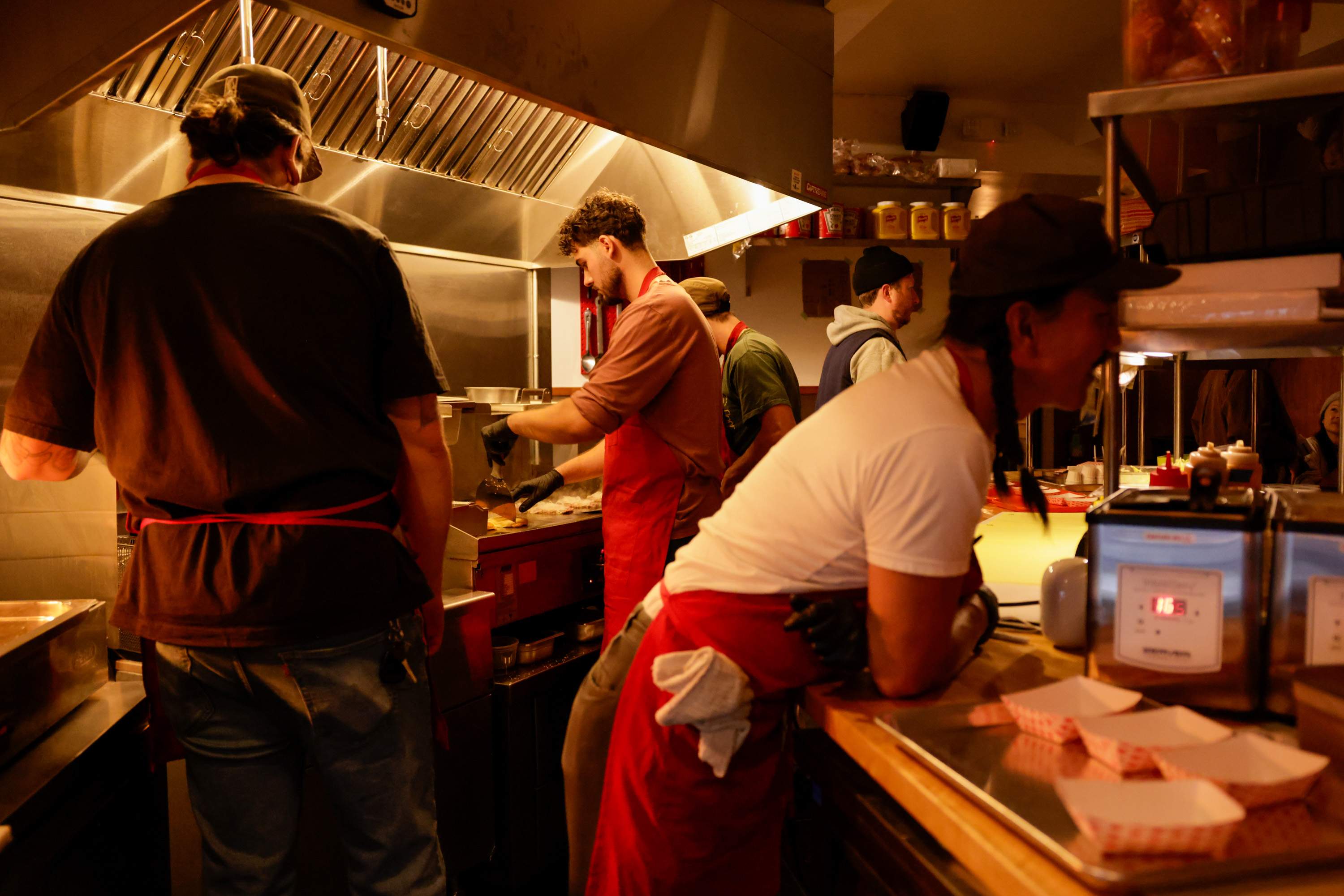 Four cooks in a bustling kitchen are preparing food. They wear aprons, and one is leaning on the counter. The space is warmly lit and active.