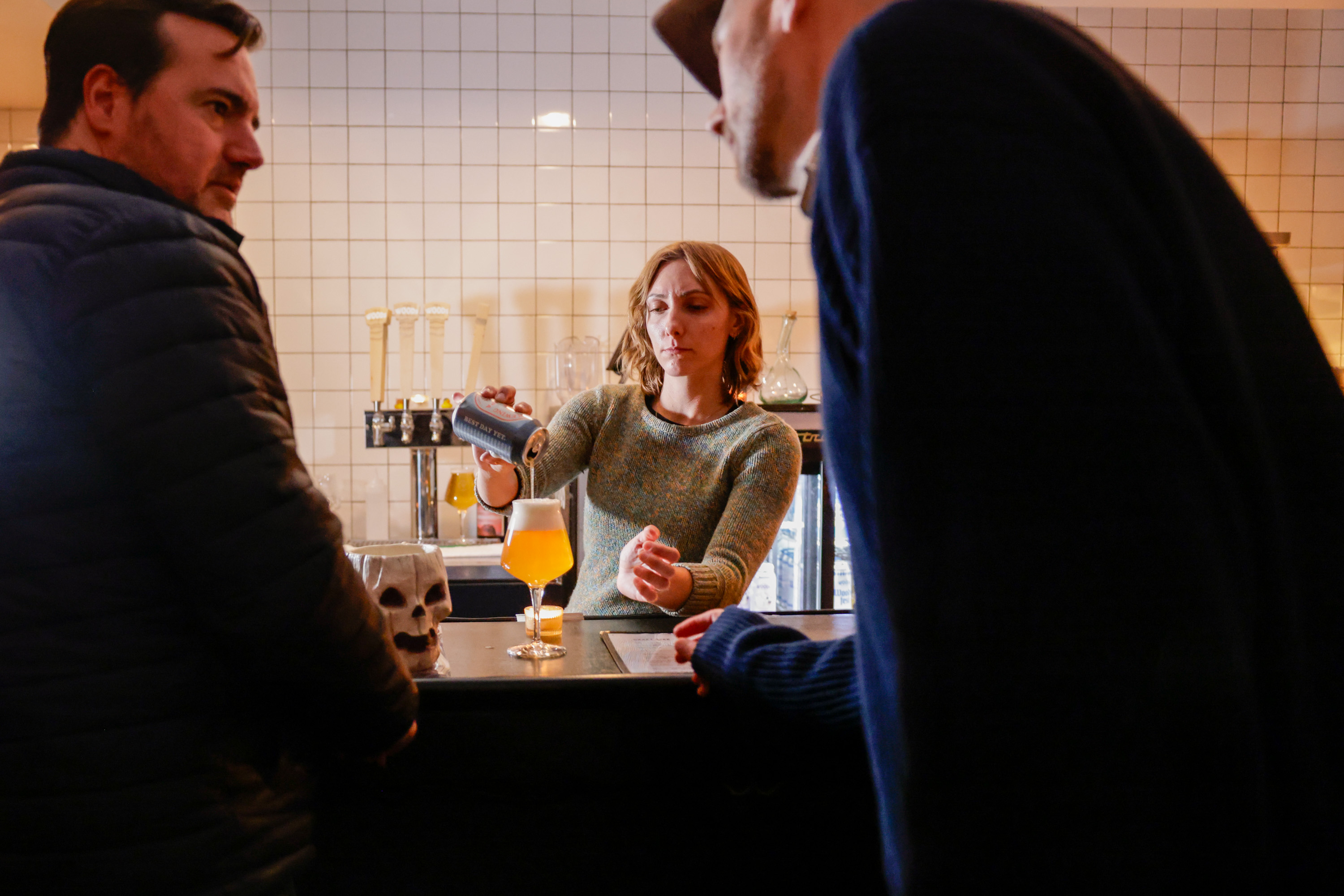 A woman pours an orange drink from a can into a glass at a bar, with two men nearby and a skull decoration on the counter. The setting is cozy and casual.