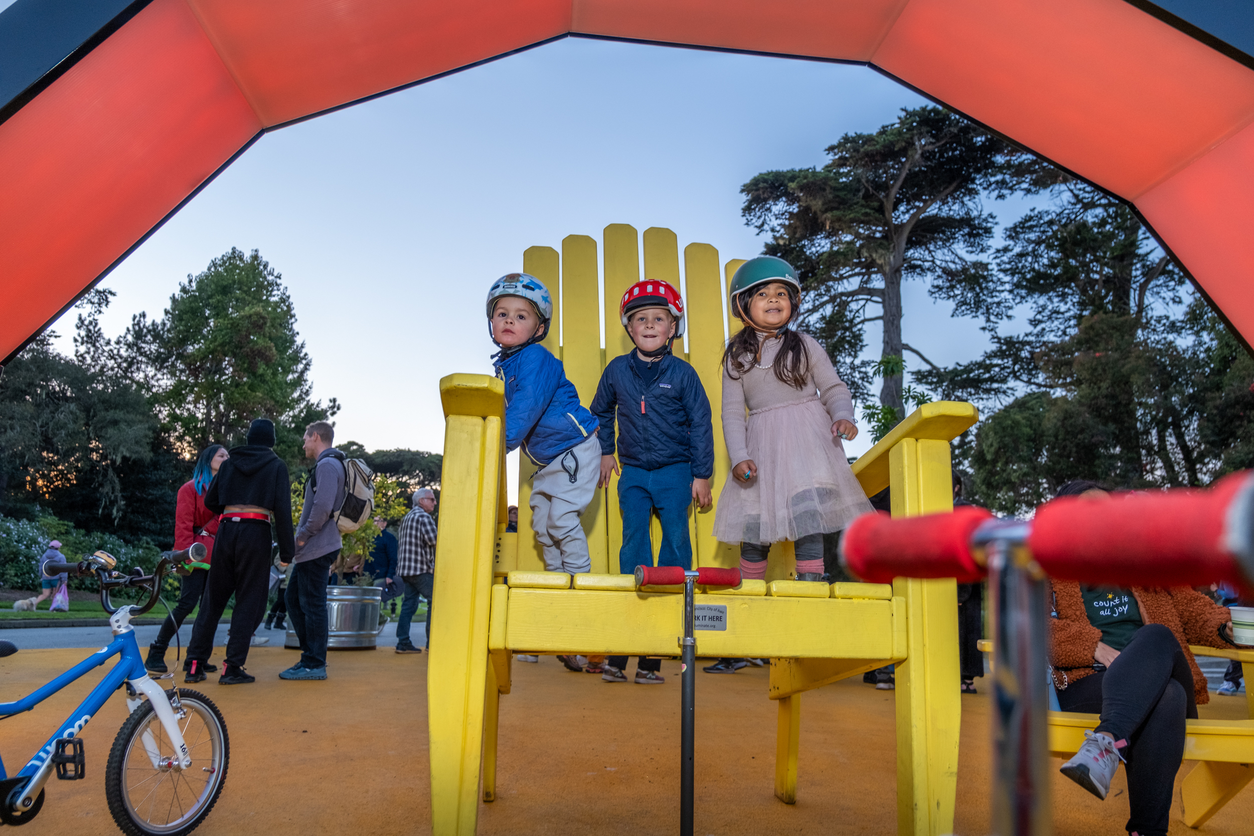 Three children wearing helmets are playing on a large yellow chair in an outdoor park setting. A blue bike is parked nearby, and several adults are gathered around.