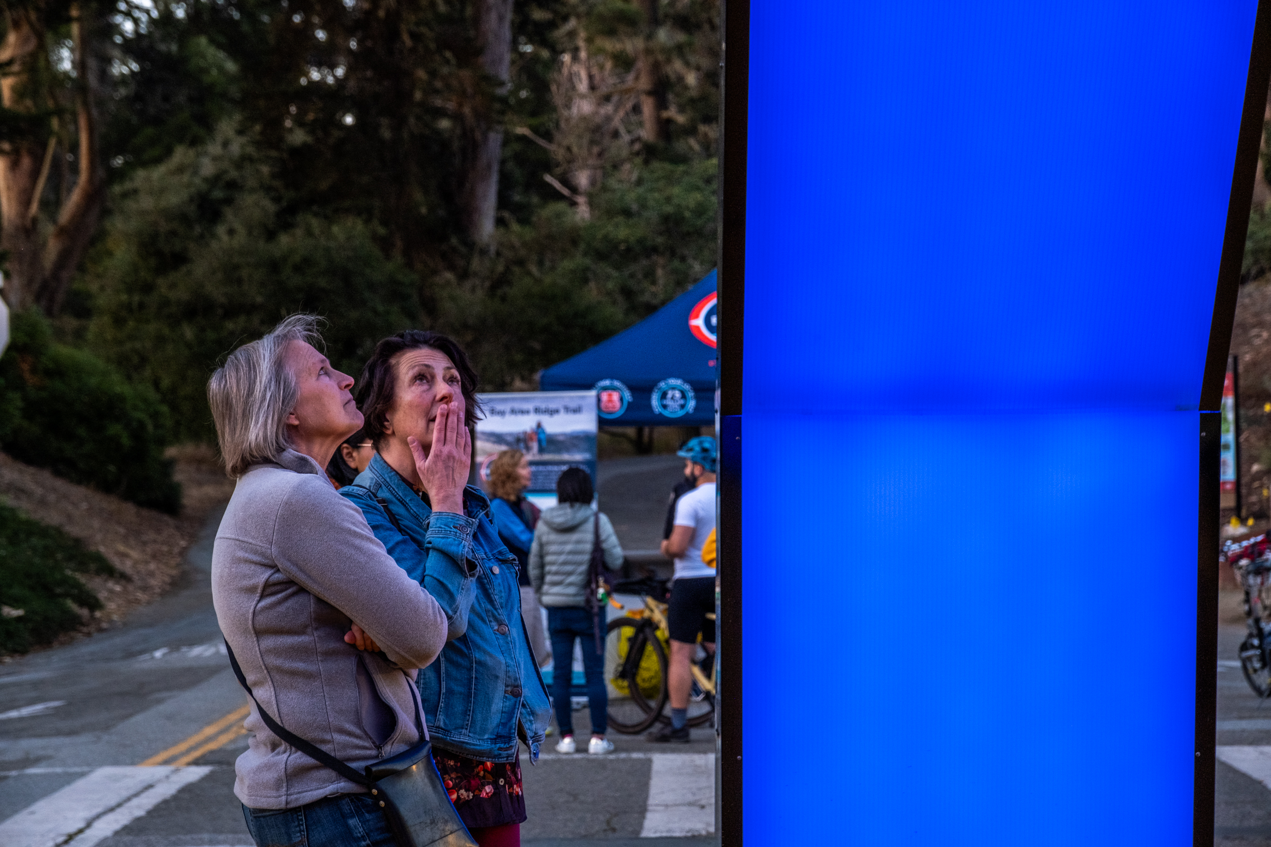 Two women stand outdoors, looking intently at a large, illuminated blue screen. In the background, people gather near a tent and trees.