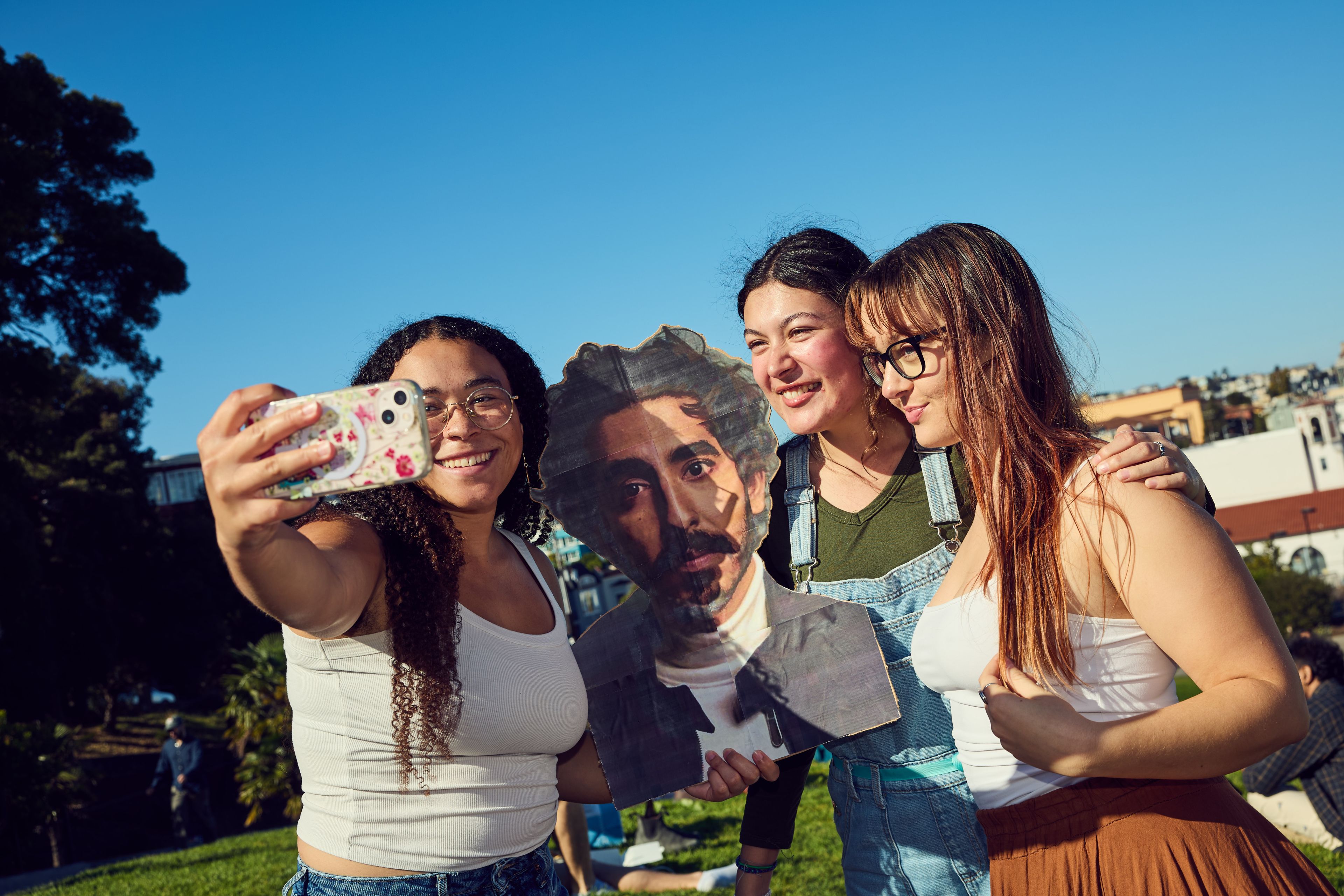 Three people pose for a selfie on a sunny day with a cardboard cutout of a man. They are outdoors on grassy terrain, smiling joyfully under a clear blue sky.