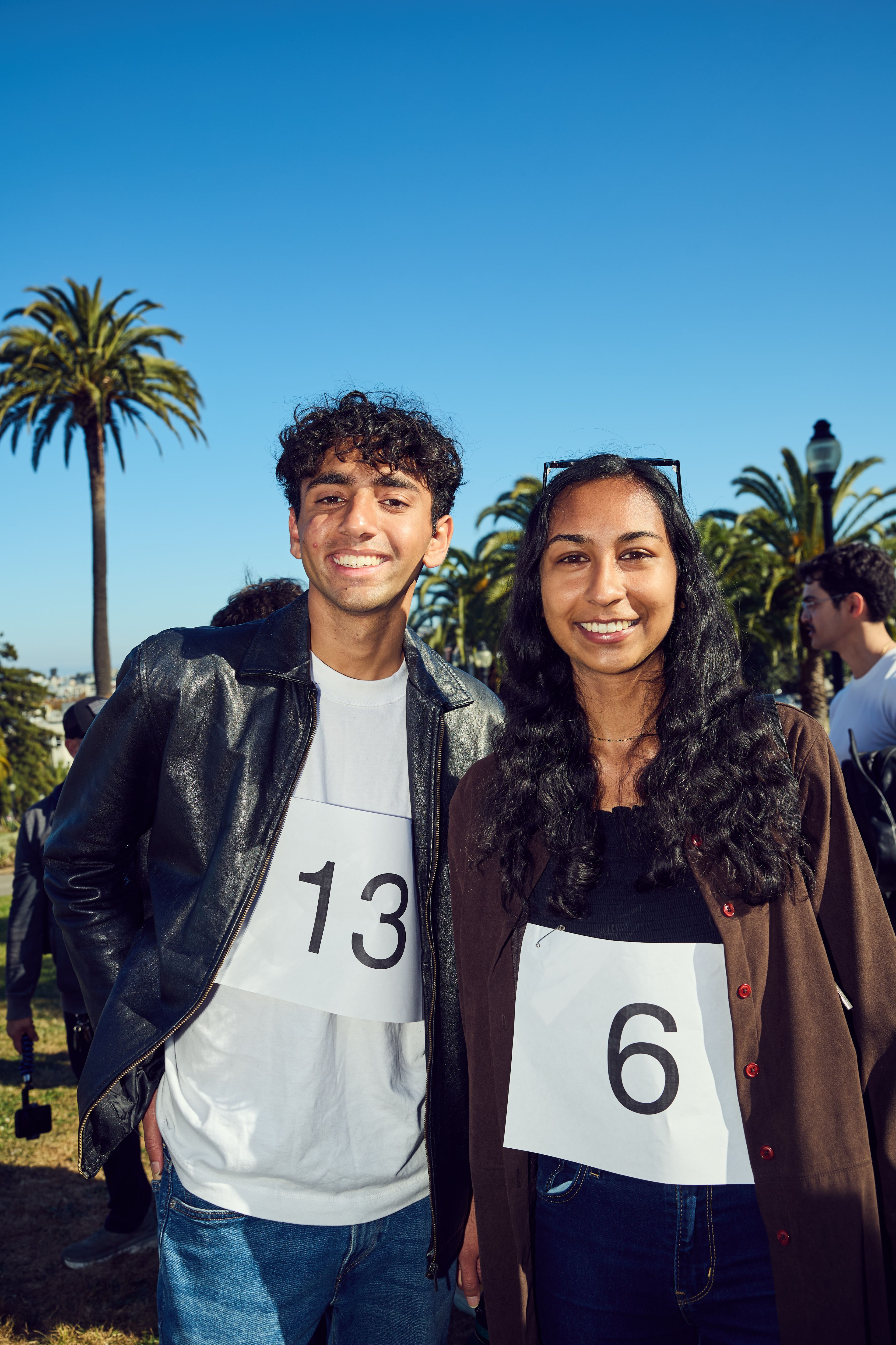 Two people stand smiling outdoors, wearing numbered papers &quot;13&quot; and &quot;6&quot;. Palm trees and a clear blue sky fill the background.