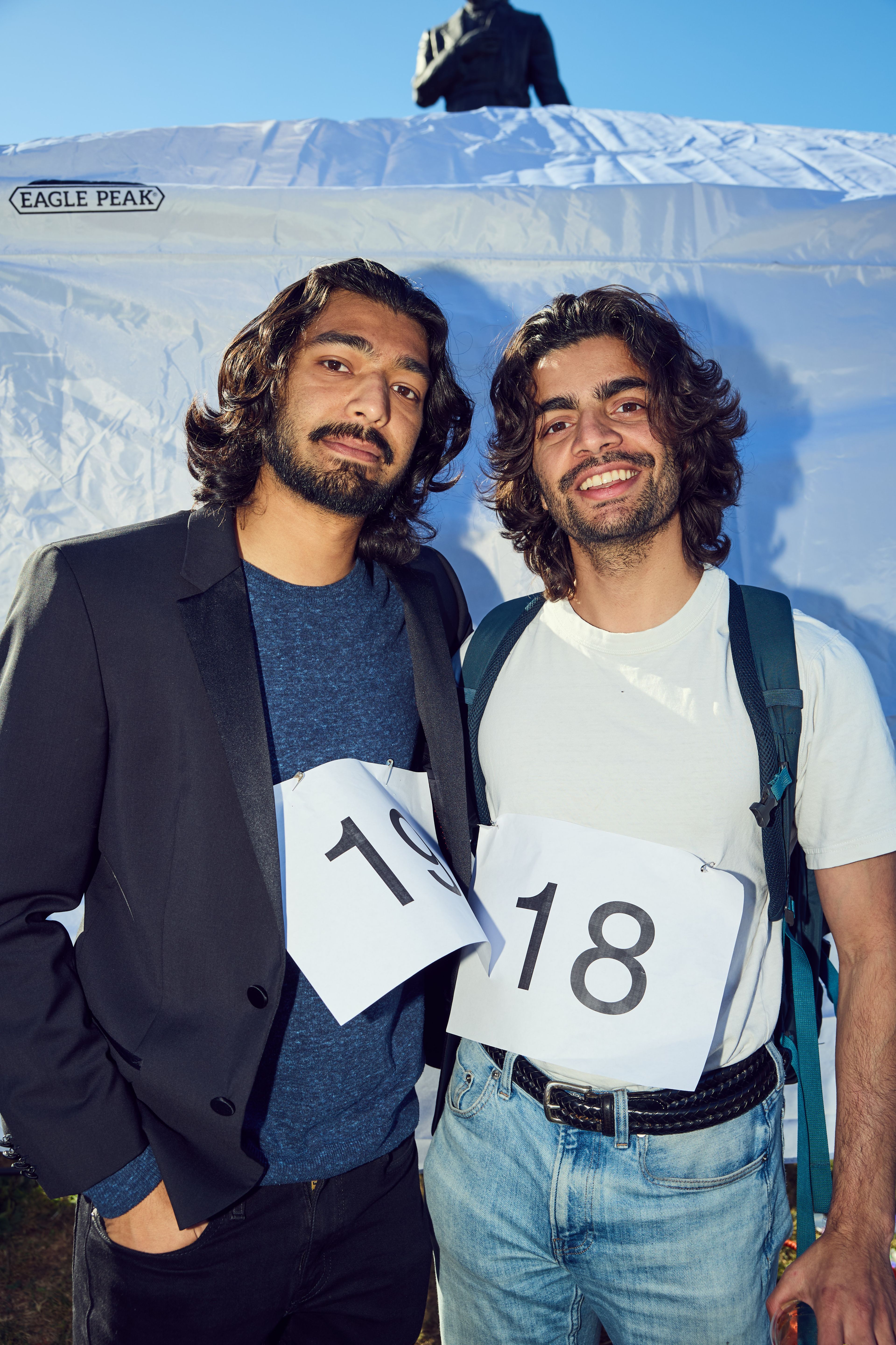 Two men with long hair and beards stand smiling. One wears a blazer and dark shirt, the other a white t-shirt and backpack. They have numbers 19 and 18 on paper.