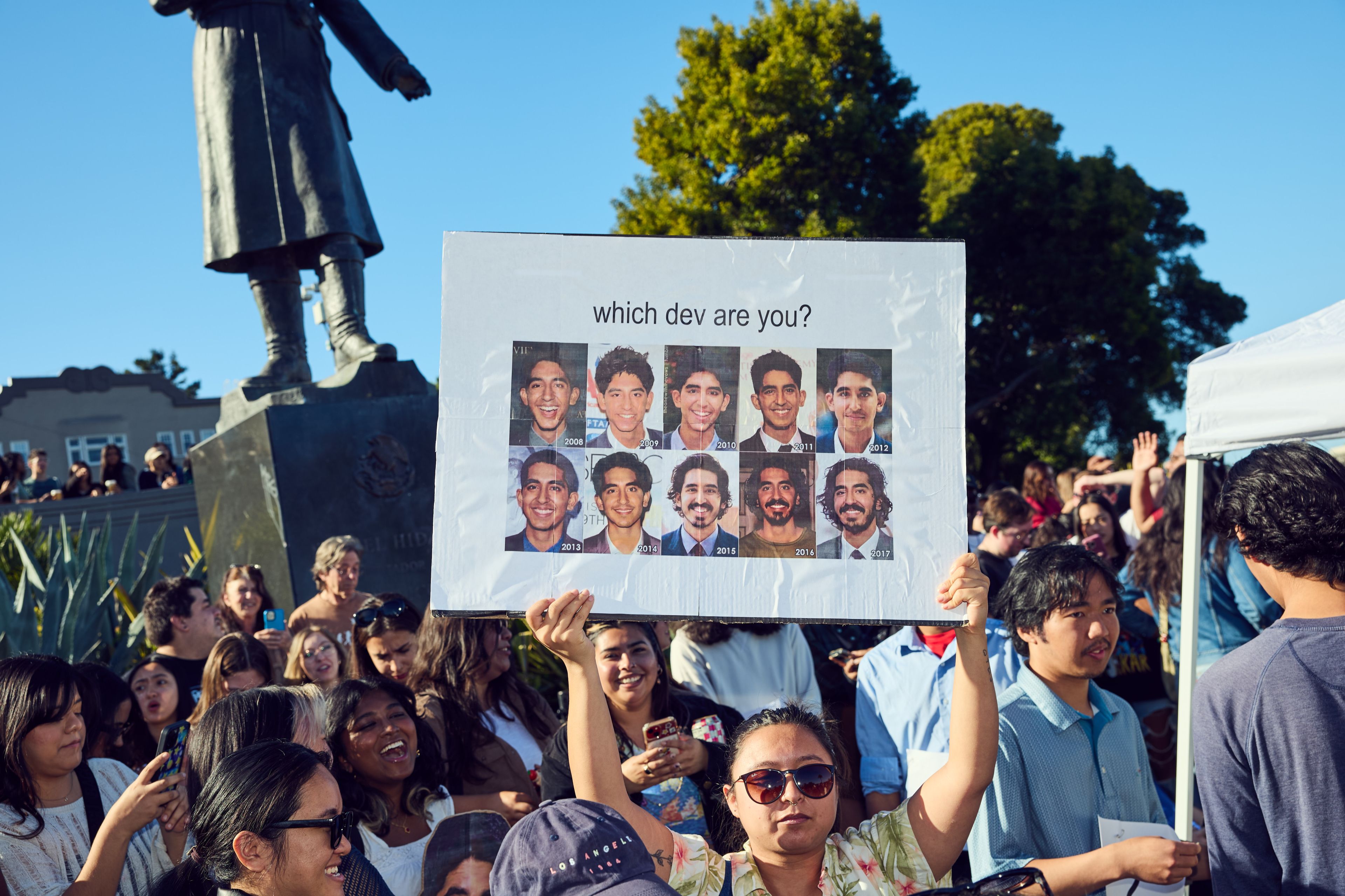 A person holds a sign with various years and faces, asking &quot;which dev are you?&quot; amid a crowd near a large statue, under a clear blue sky.