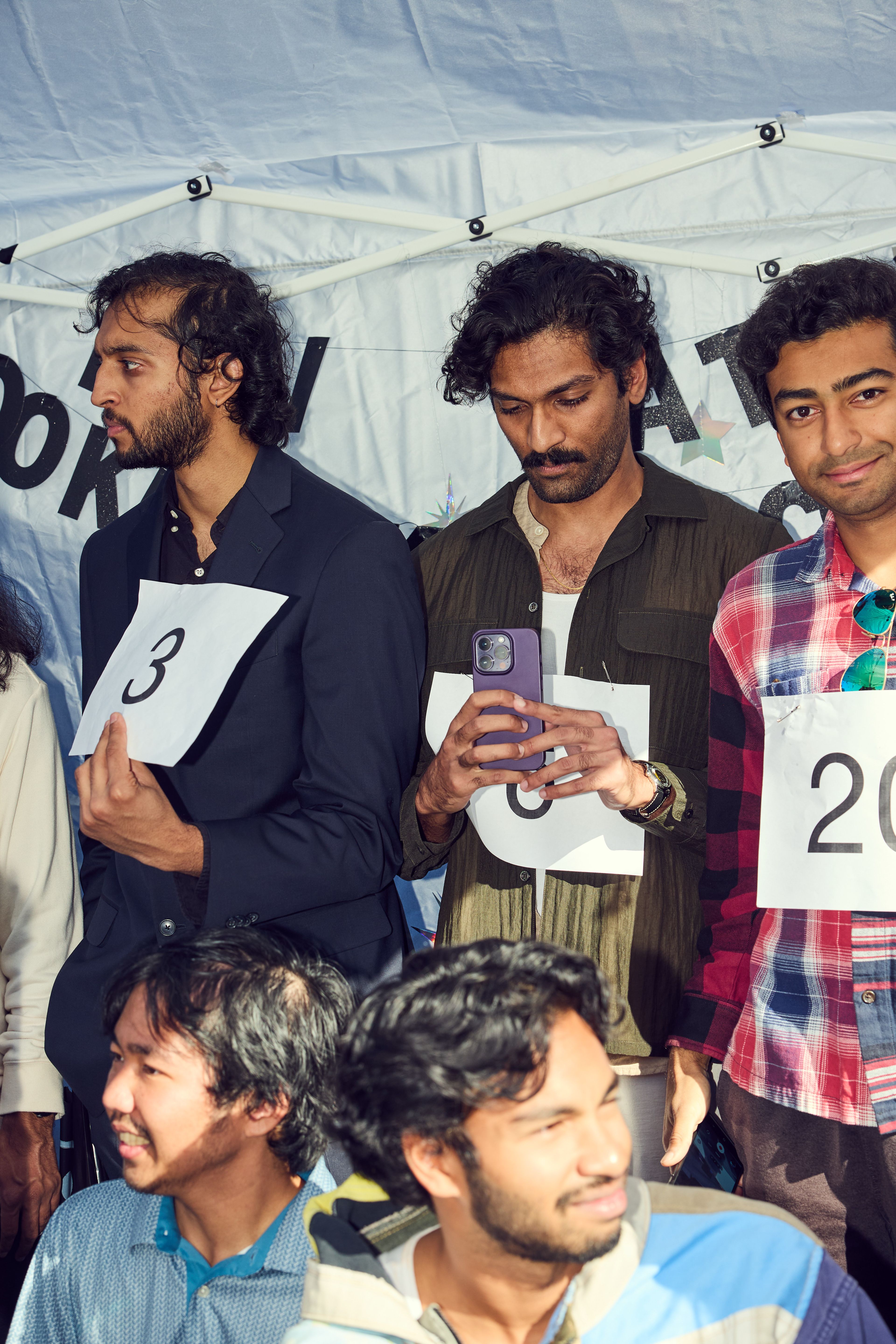 Five people are in a group, each wearing numbered paper signs. One person holds a smartphone, and they are under a white tent with letters partially visible.