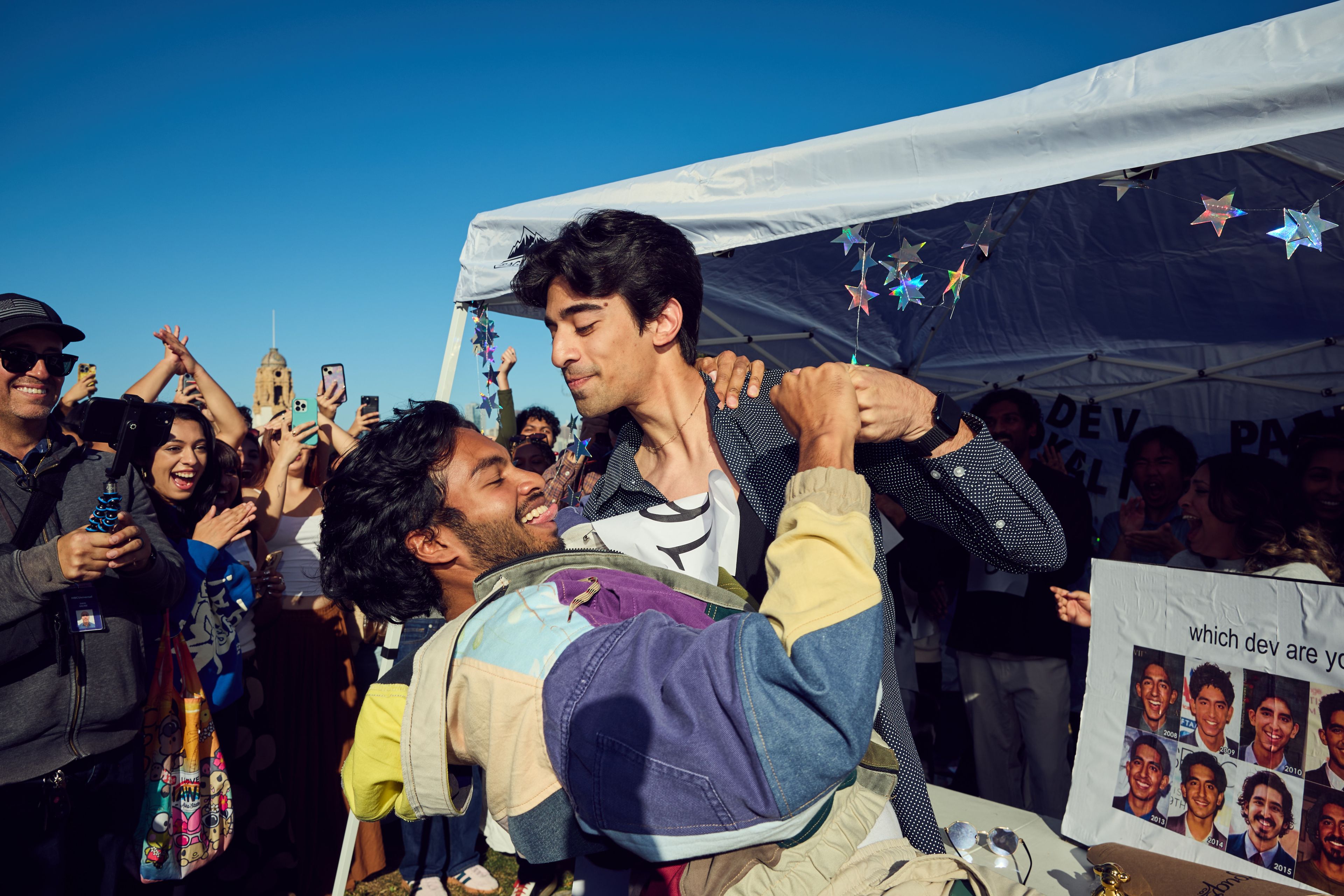 Two people share a playful moment in a lively, outdoor gathering. One person is dipping the other, while a joyous crowd with phones cheers them on, under a tent adorned with stars.