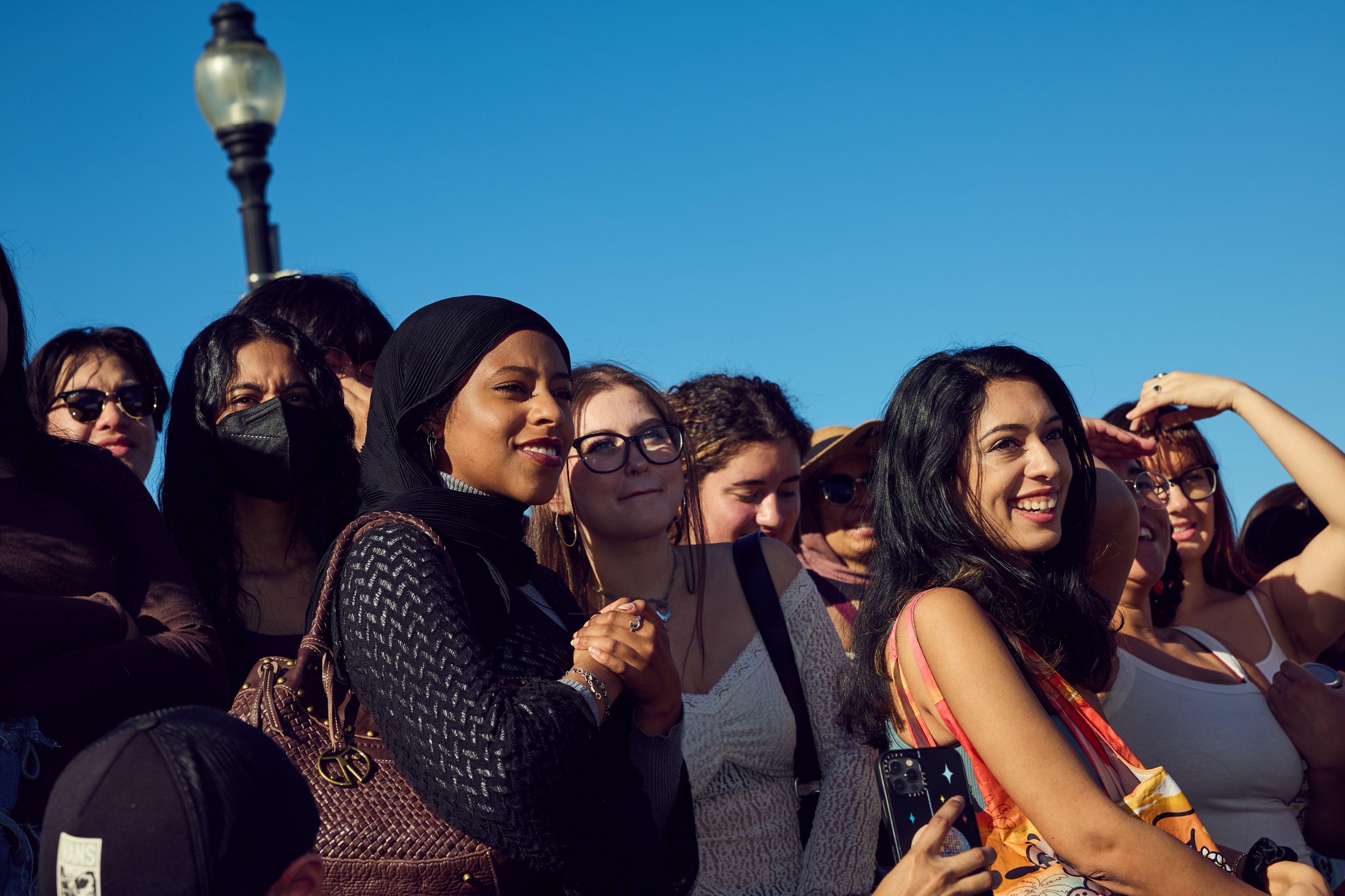 A diverse group of people, mostly women, are gathered outside under a clear blue sky. They are smiling and appear to be enjoying an event, with a streetlamp in the background.