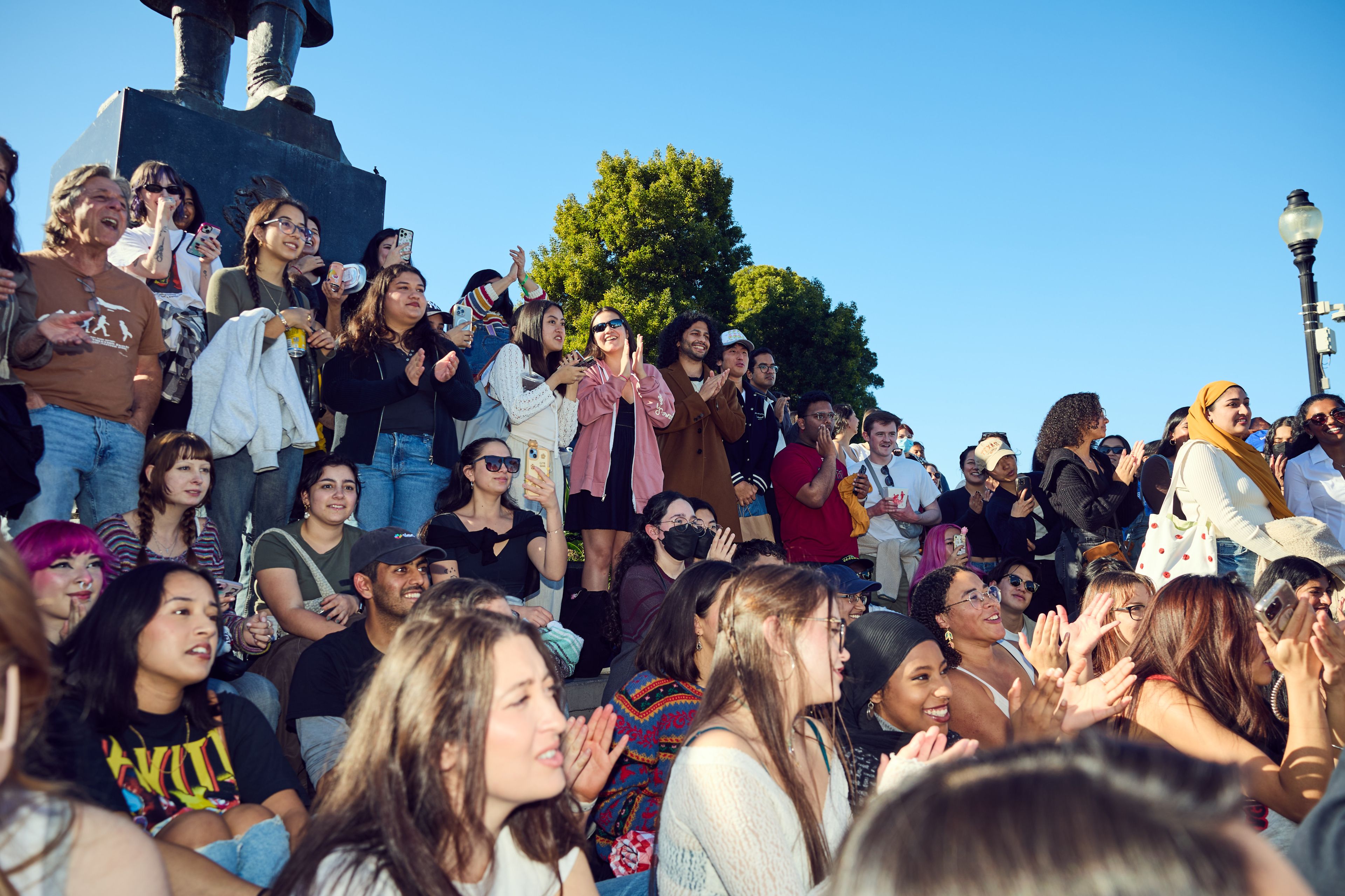 A diverse group of people is gathered outdoors in bright daylight, clapping and smiling. They are seated and standing, some holding phones, against a backdrop of trees and a blue sky.