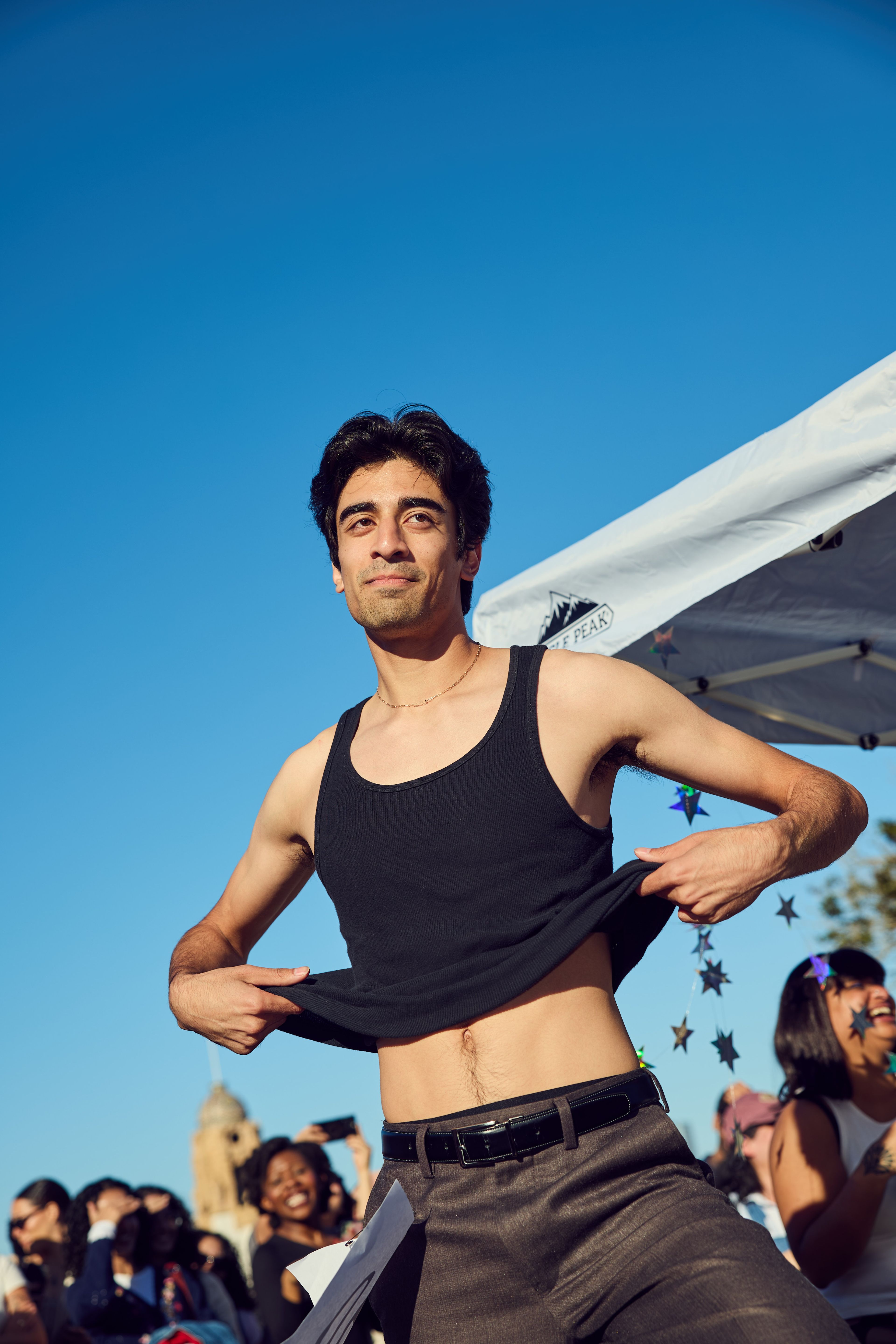 A person in a black tank top is outdoors, pulling up their shirt slightly. It's sunny, with a crowd in the background, and a canopy overhead.