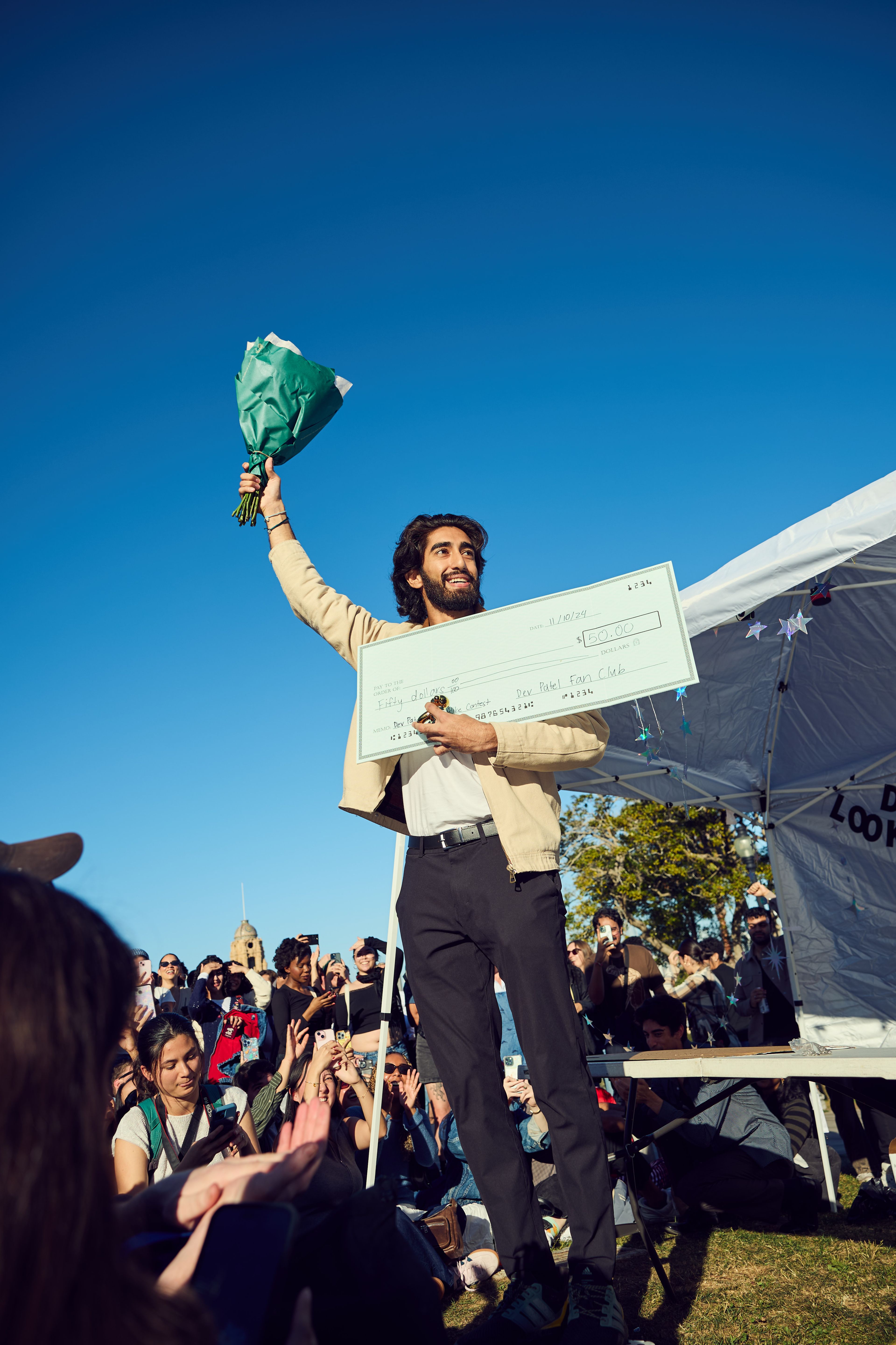 A man stands outdoors, holding a bouquet and a large check, smiling at a crowd. People around him are clapping and taking photos against a clear blue sky.