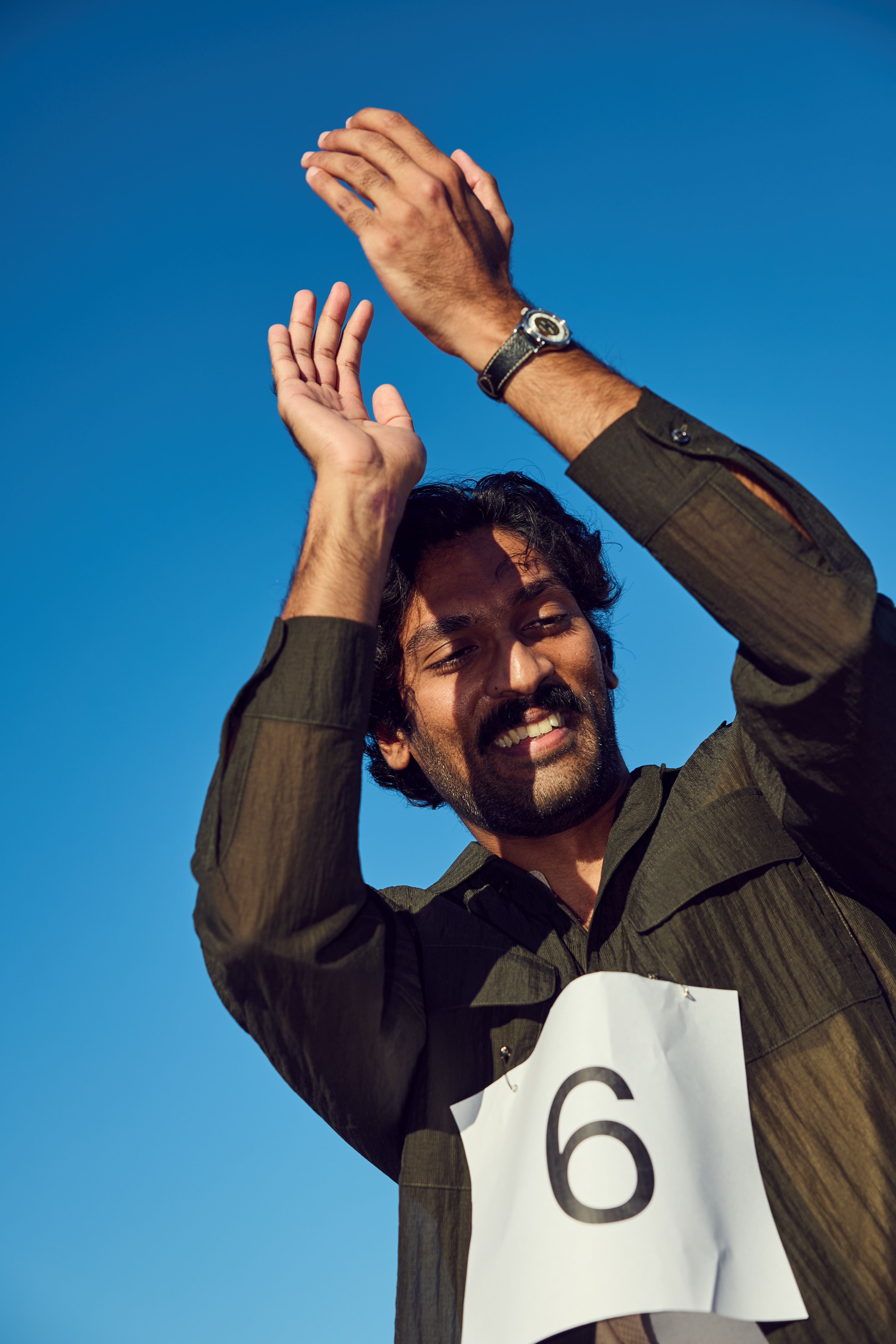 A smiling man with a mustache, dressed in a dark shirt with a number &quot;6&quot; pinned on it, is clapping under a clear blue sky.