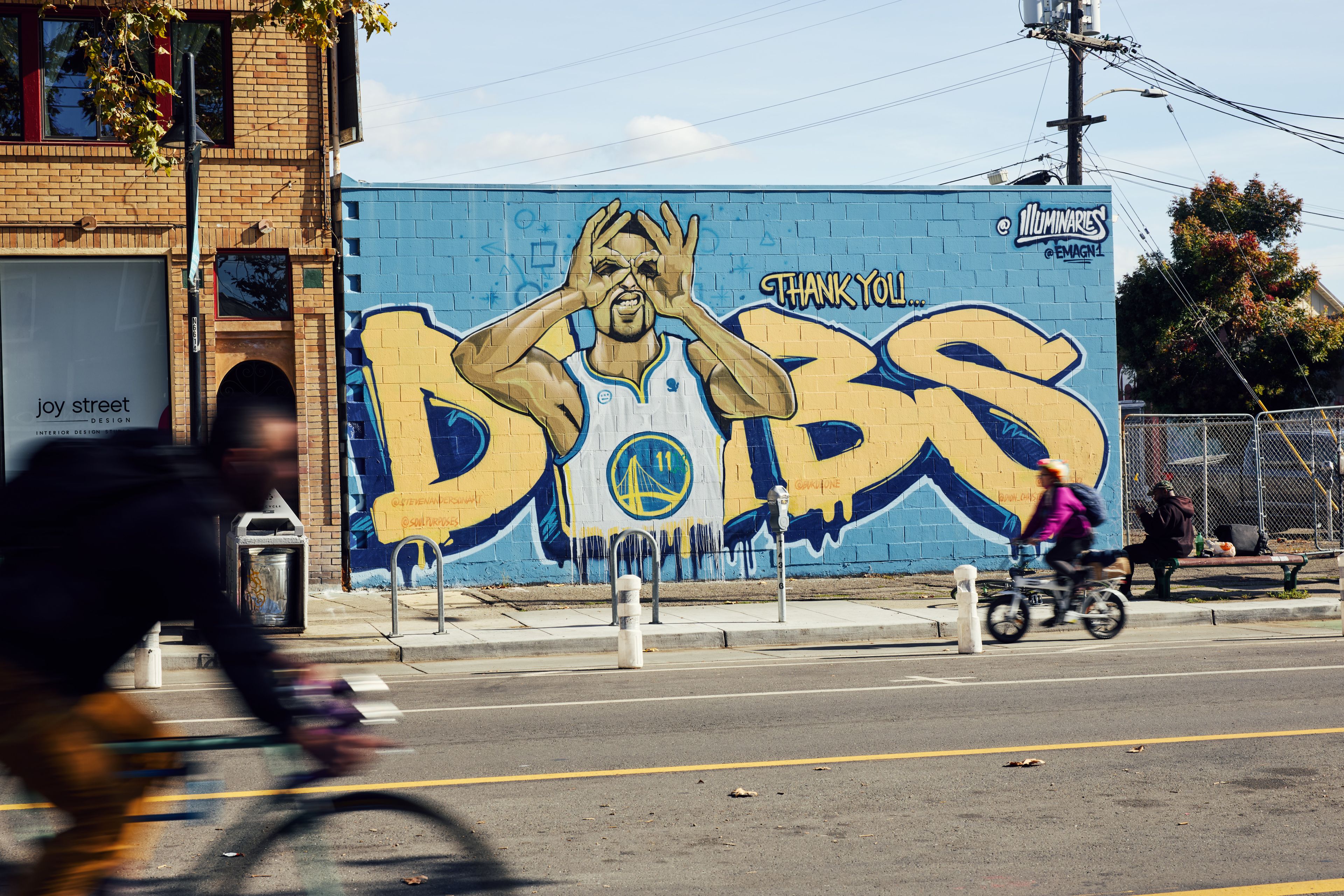 A colorful mural features a basketball player in a white jersey with large graffiti-style letters spelling &quot;DUBS&quot; and a &quot;Thank you&quot; message. Two cyclists pass by in motion.