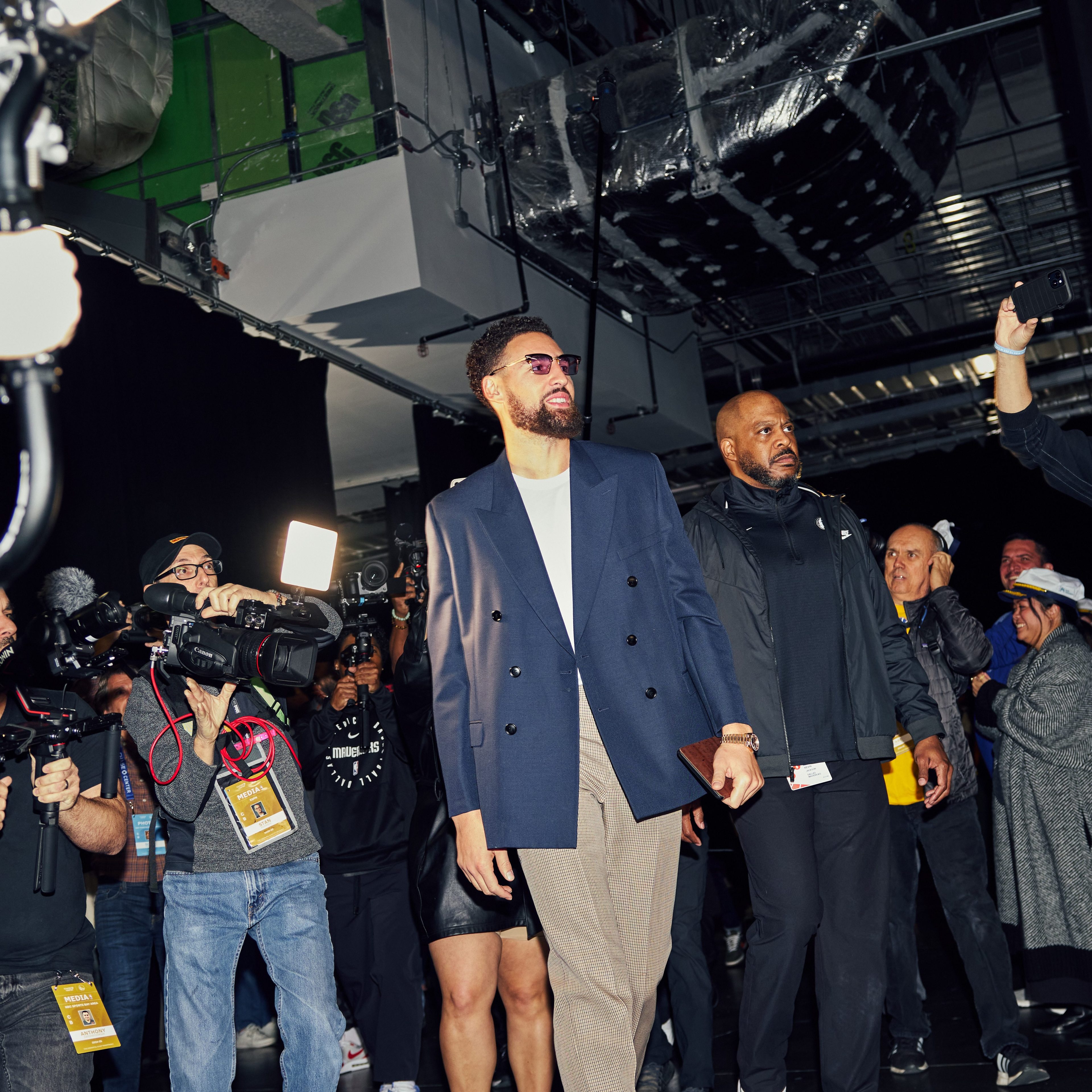 A well-dressed man walks surrounded by photographers and media, capturing his presence in a crowded, industrial-like setting.