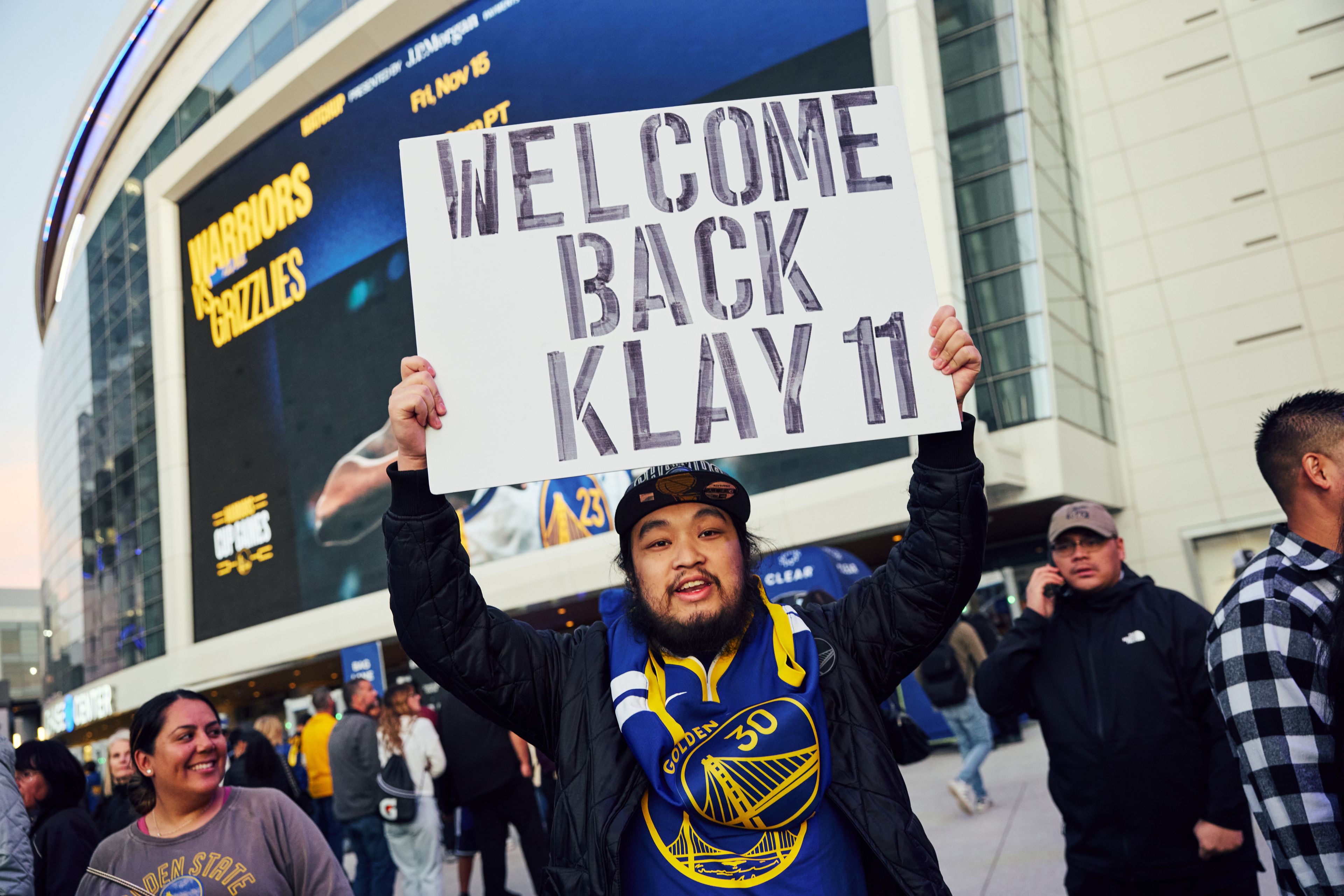 A fan outside a sports arena holds a sign reading &quot;WELCOME BACK KLAY 11,&quot; wearing team gear, amidst a crowd. The building displays &quot;WARRIORS vs GRIZZLIES.&quot;