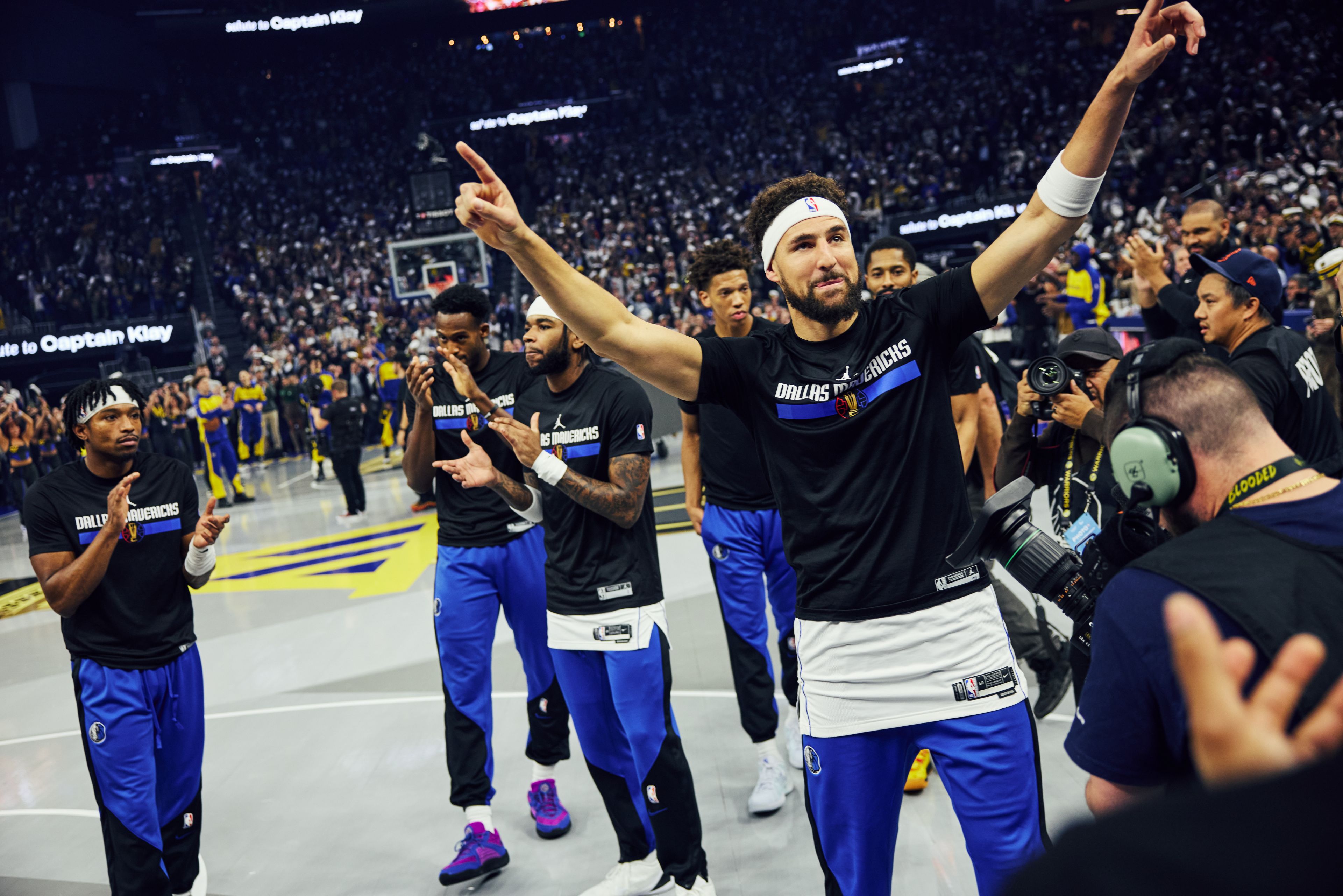 Basketball players from the Dallas Mavericks celebrate on the court amid a cheering crowd and photographers. One player gestures with his hand raised.