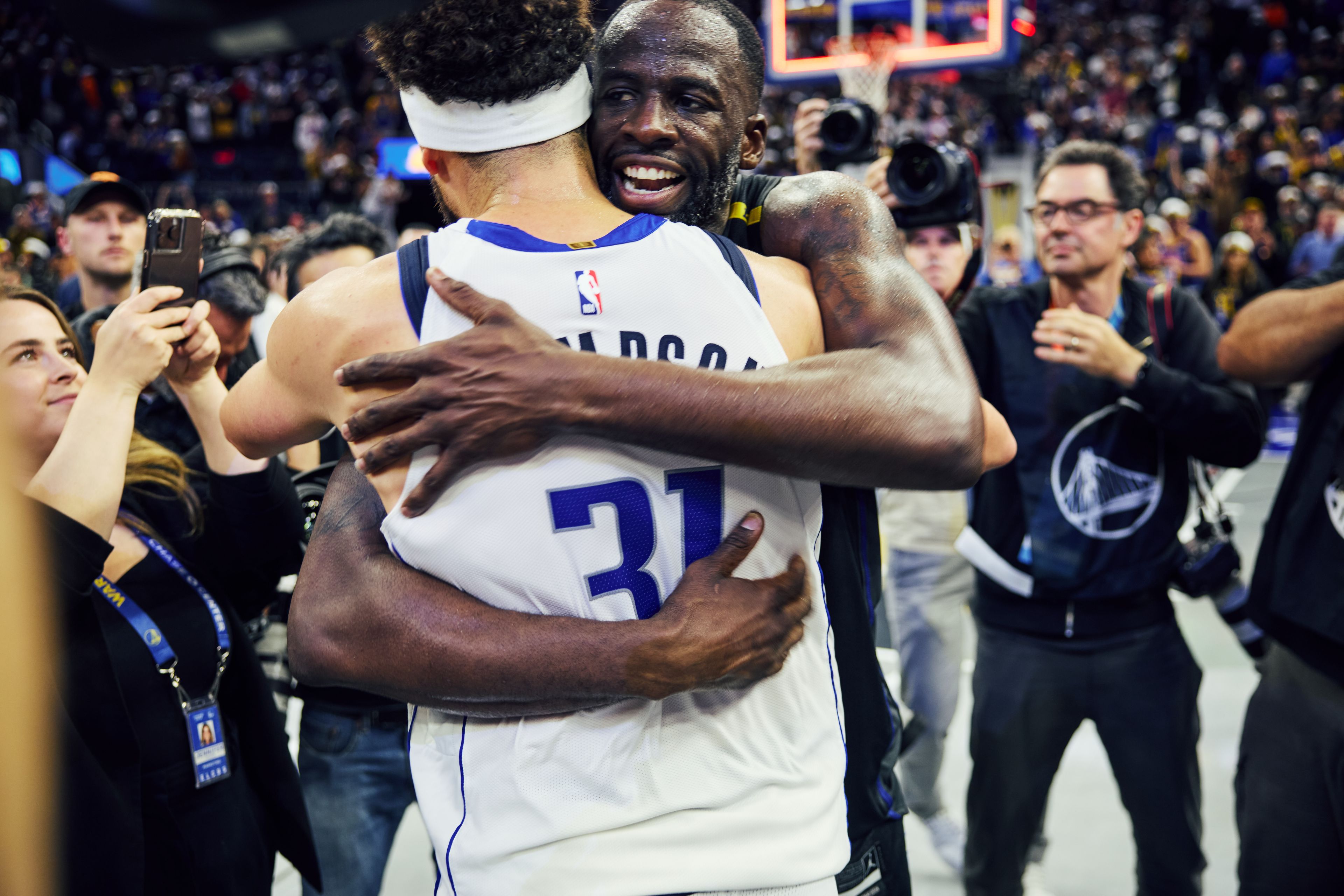 Two basketball players are hugging on the court while surrounded by photographers and fans. One player is wearing a headband and a white jersey.