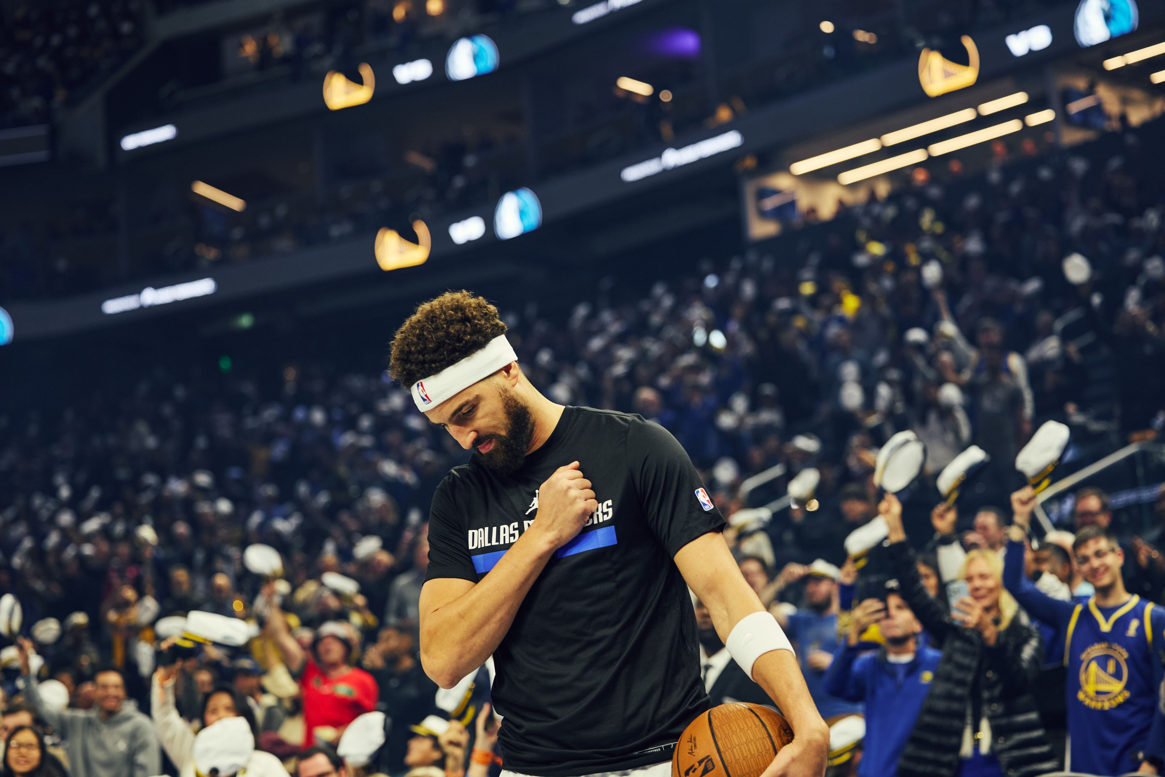 A basketball player in a "Dallas Mavericks" shirt stands on the court, holding a ball, with a cheering crowd in the background. He appears focused and introspective.