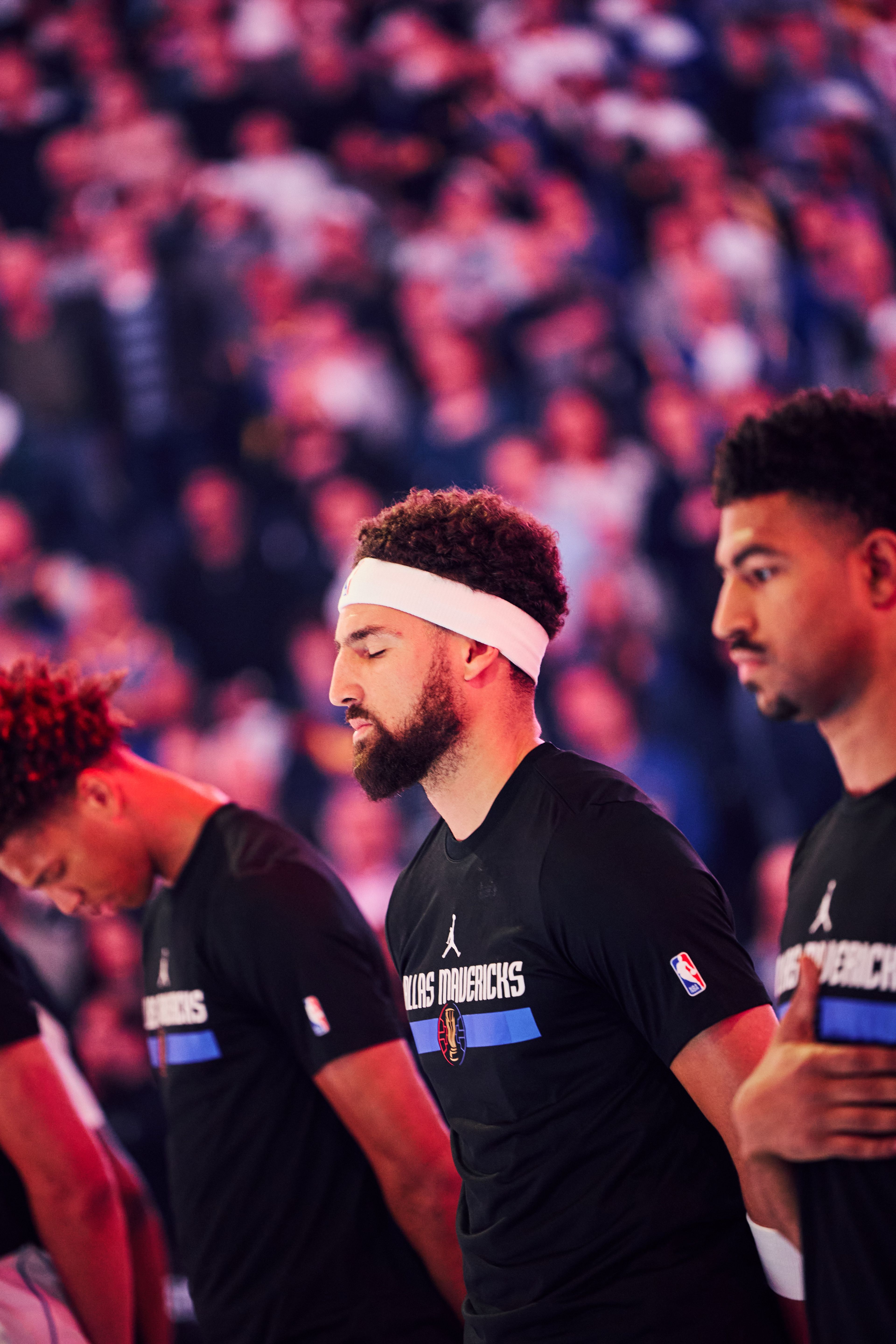 Three basketball players stand with bowed heads, wearing Dallas Mavericks shirts. The background shows a blurred crowd, creating a focused and solemn scene.