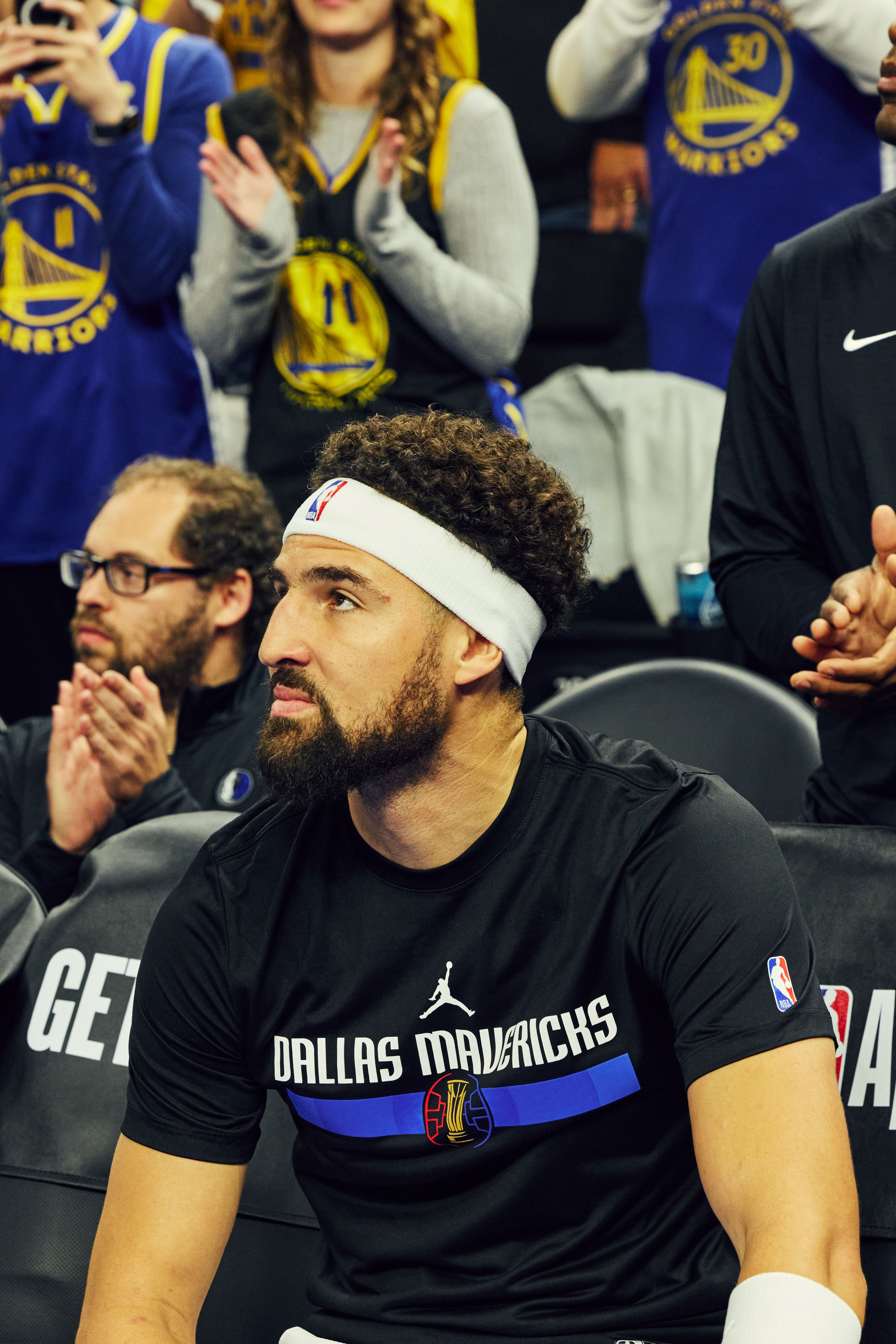 A basketball player wearing a headband and a Dallas Mavericks shirt sits on the bench, while fans in Golden State Warriors jerseys applaud behind him.