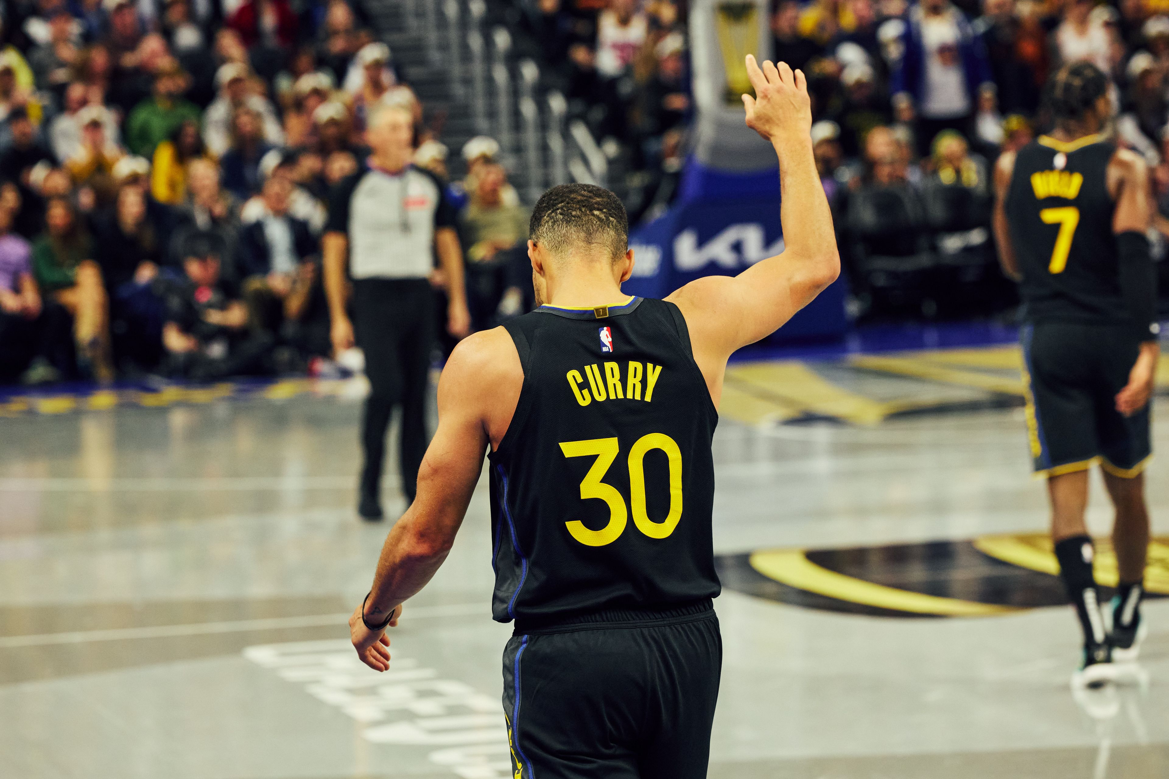 A basketball player in a black jersey with the number 30 and the name "Curry" waves his hand on the court during a game, with a crowd in the background.