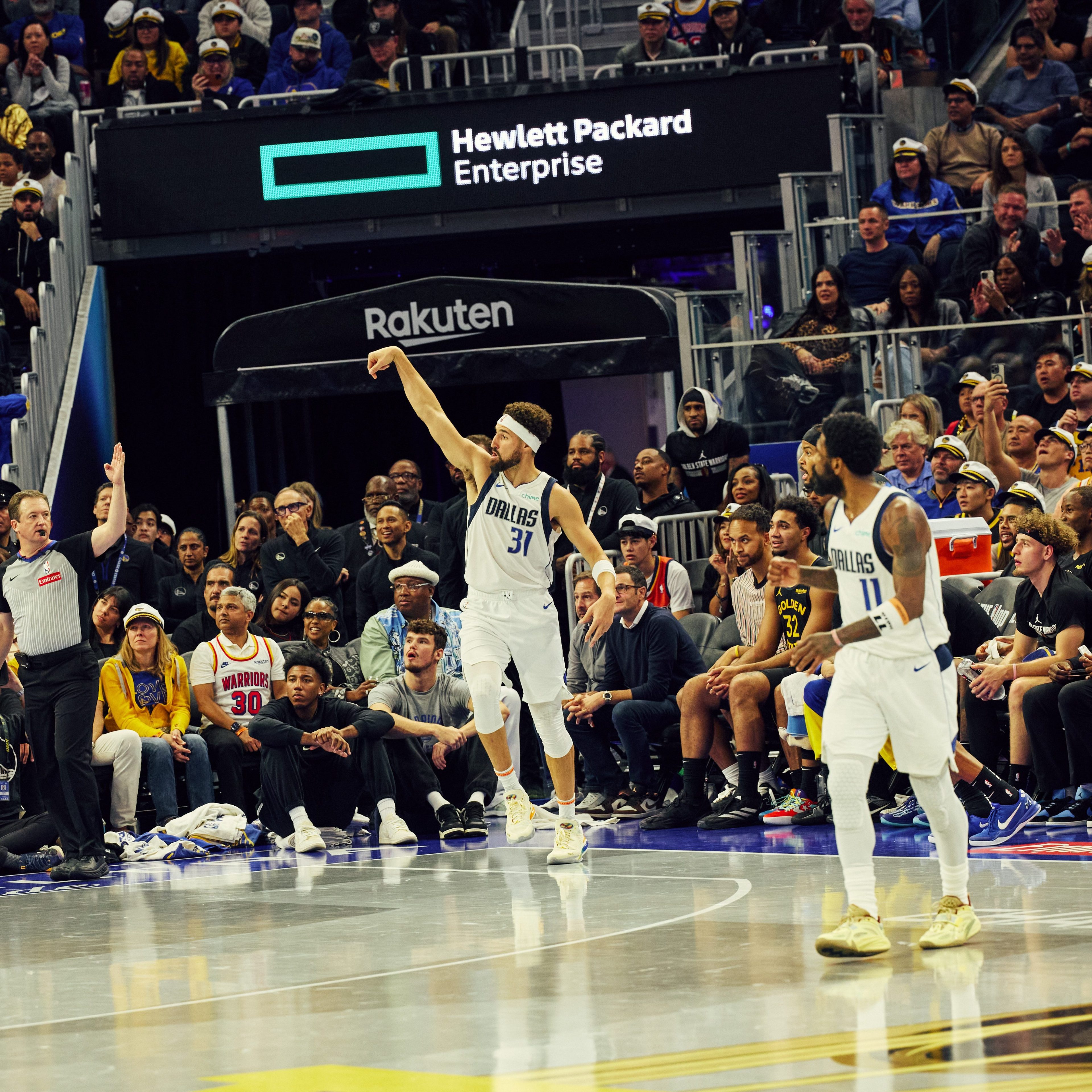 A basketball player in a white Dallas jersey is executing a jump shot, while a referee signals, surrounded by a lively crowd and other players on the court.