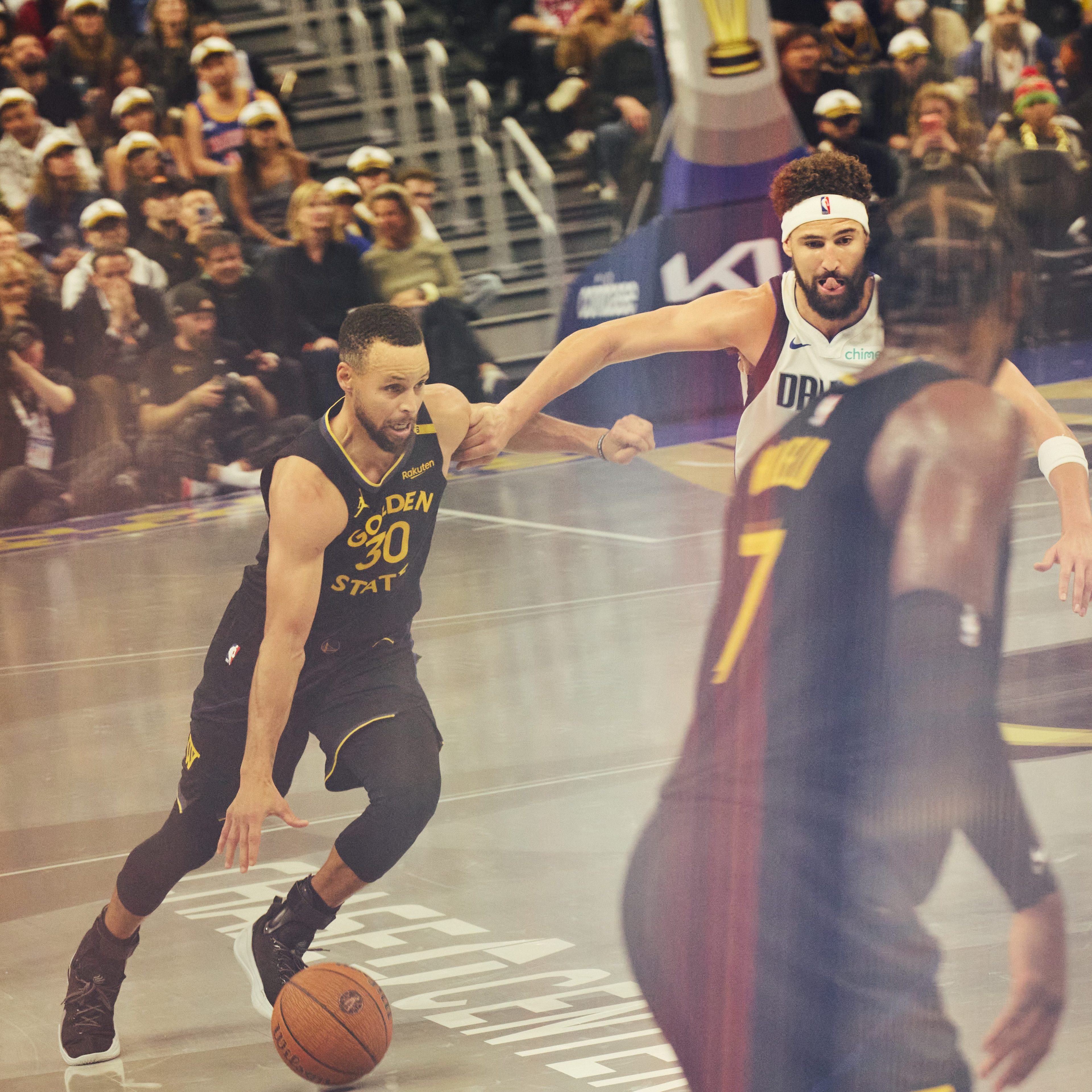 A basketball player in a black &quot;Golden State&quot; jersey dribbles the ball towards the basket while being pursued by another player in a white jersey, with a referee nearby.