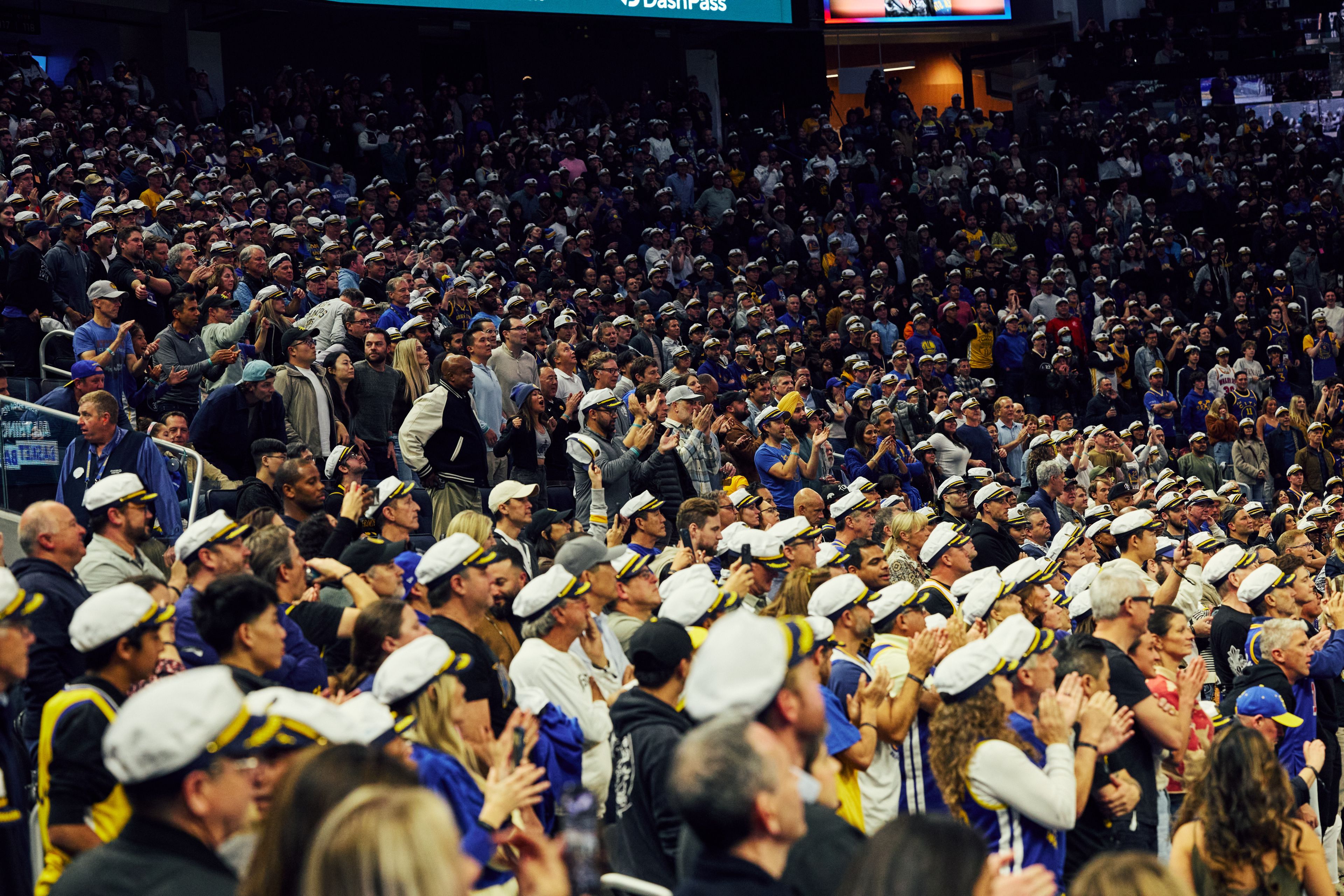 A crowd of enthusiastic fans, many wearing sailor hats, are gathered in a stadium, standing and clapping, creating a lively and energetic atmosphere.