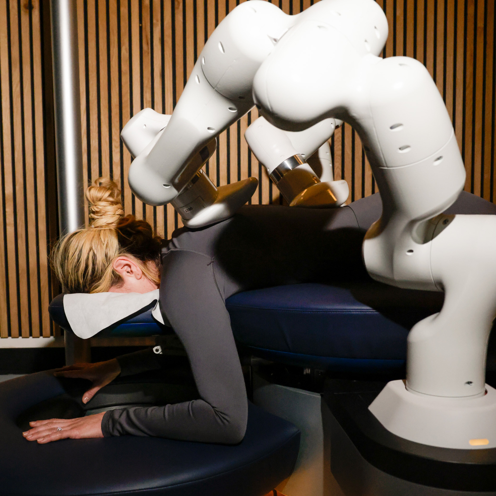 A person is lying face down on a massage table, with a robot arm hovering over their back, in a room with wooden paneling.