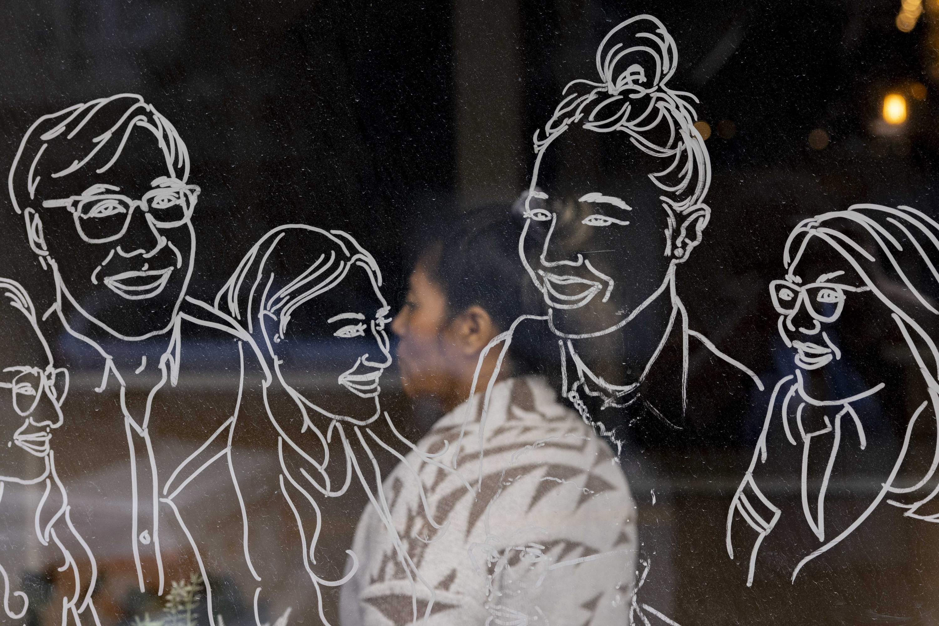 The image shows a person's reflection on a glass window adorned with white line art drawings of smiling people, creating an overlapping effect.