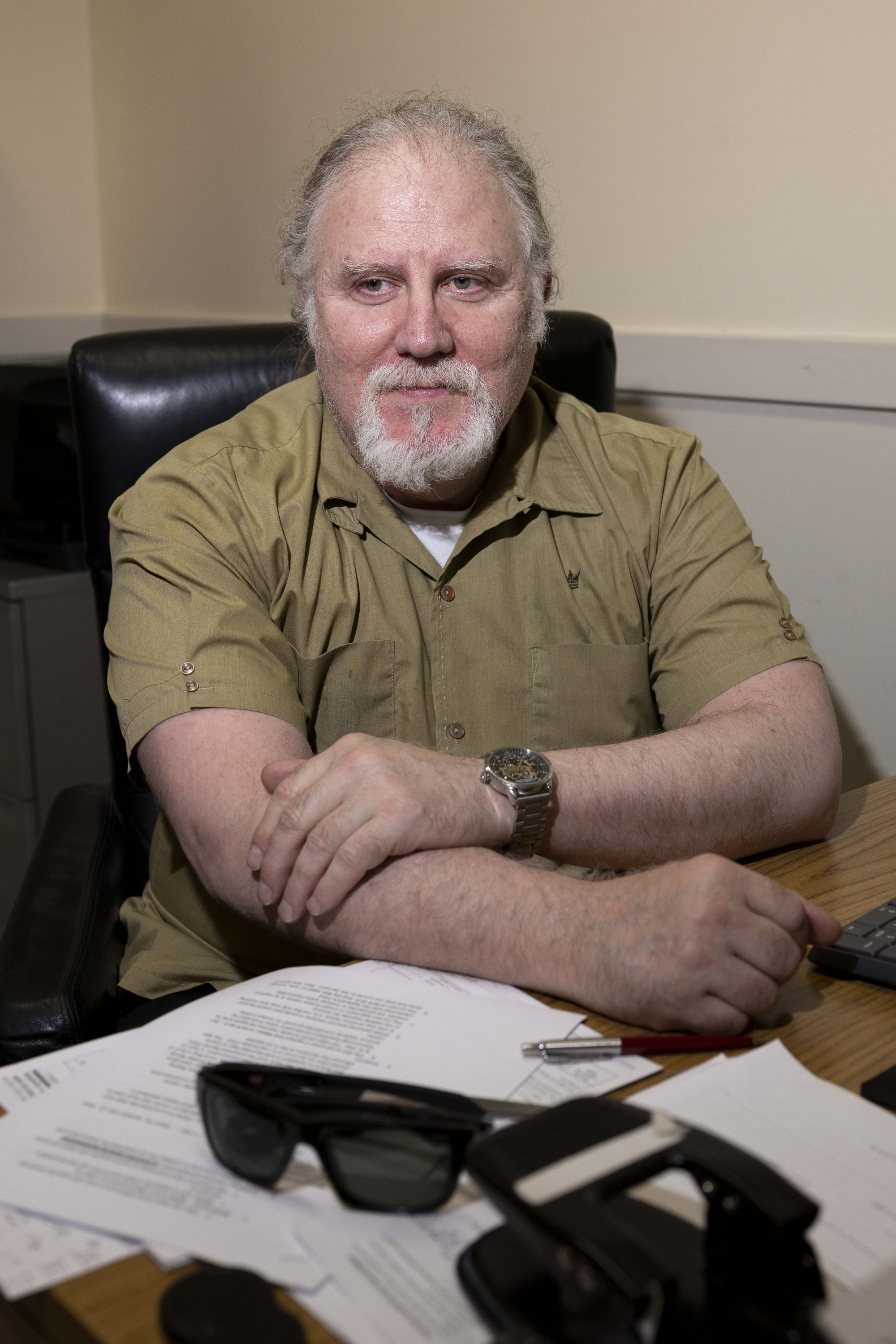 A man with a beard and mustache sits at a desk with folded arms, wearing a beige shirt and a silver watch. Papers, a pen, and sunglasses are on the desk.