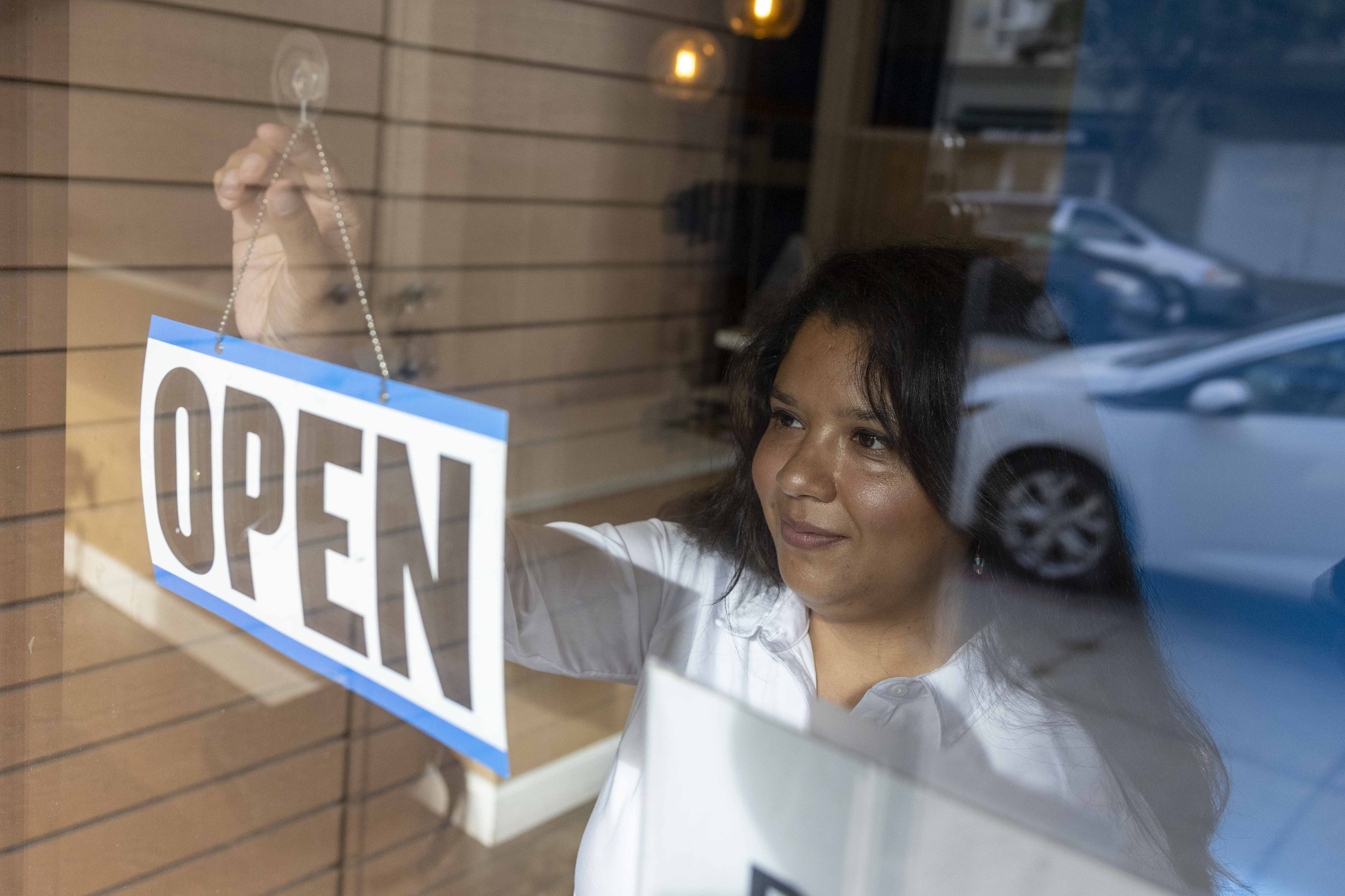 A woman is at a window, smiling as she hangs an &quot;OPEN&quot; sign. It's daytime, cars are visible outside, and warm lighting is present inside.