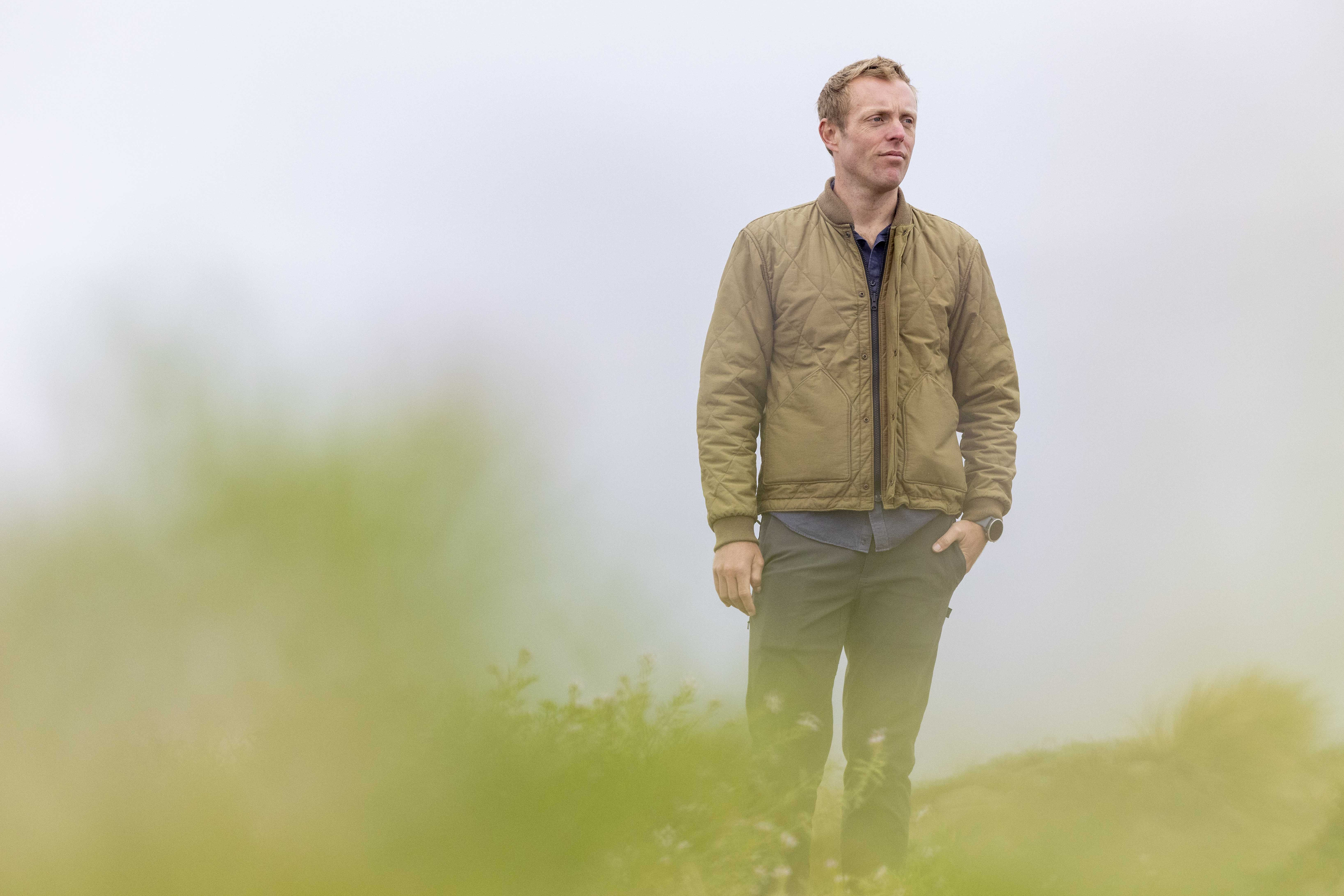 A person stands outdoors in a misty environment, wearing a brown jacket and dark pants, looking thoughtful with hands partly in pockets.