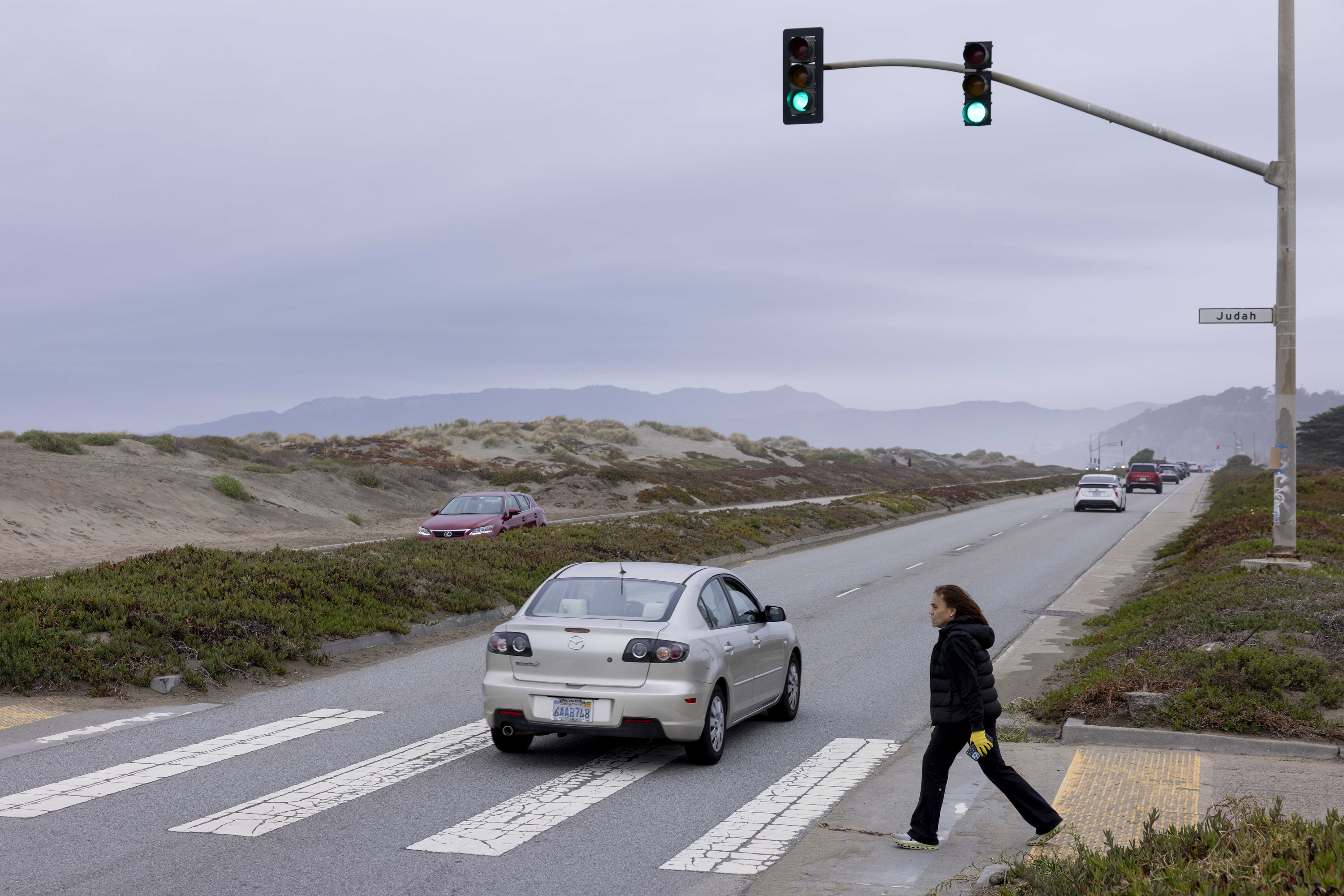 A woman crosses a street with a crosswalk, while cars pass by. A traffic light shows green, and dunes and mountains are in the background.