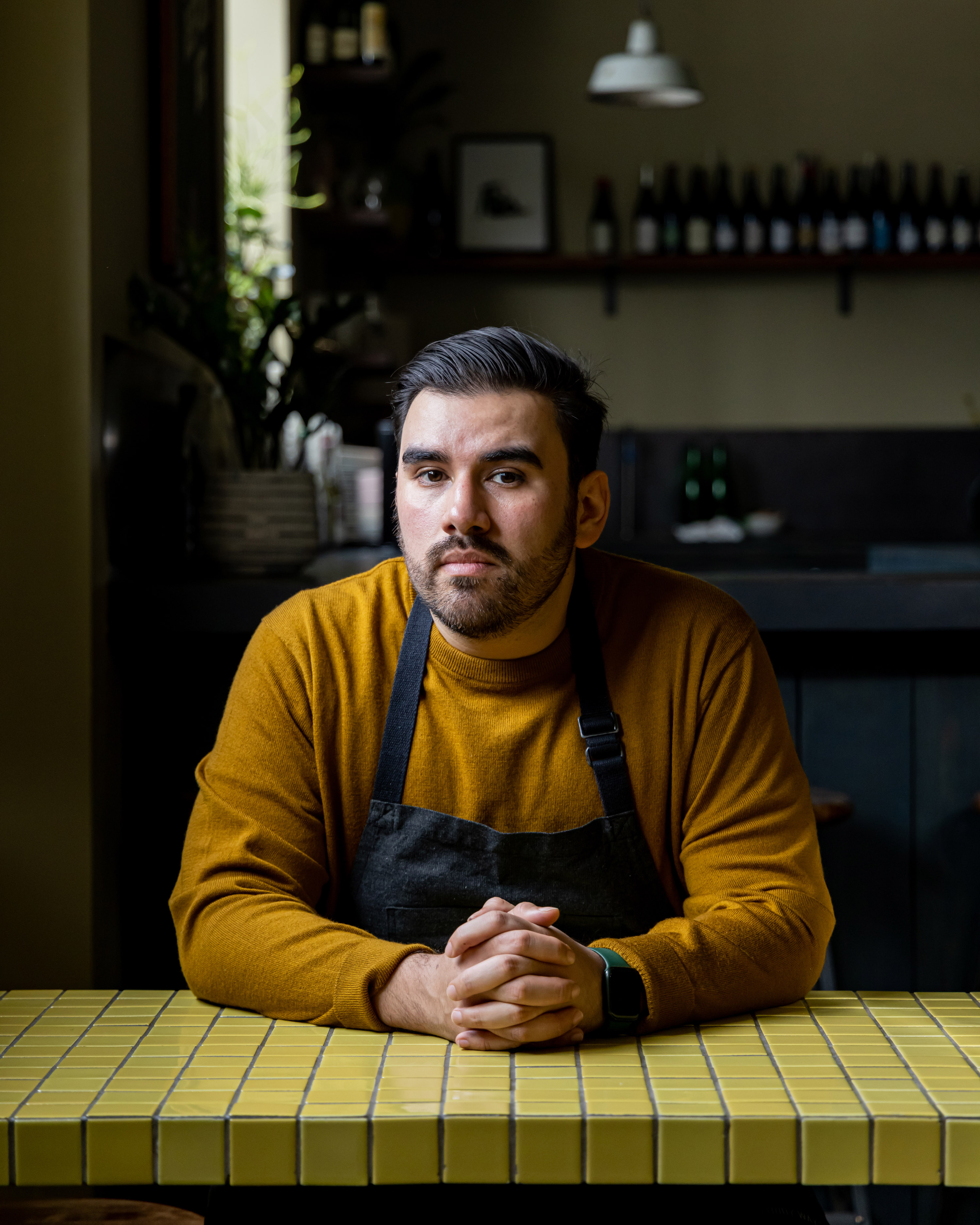 A man in a mustard sweater and apron sits at a yellow tiled table with hands clasped, in a dimly lit room with shelves and plants in the background.