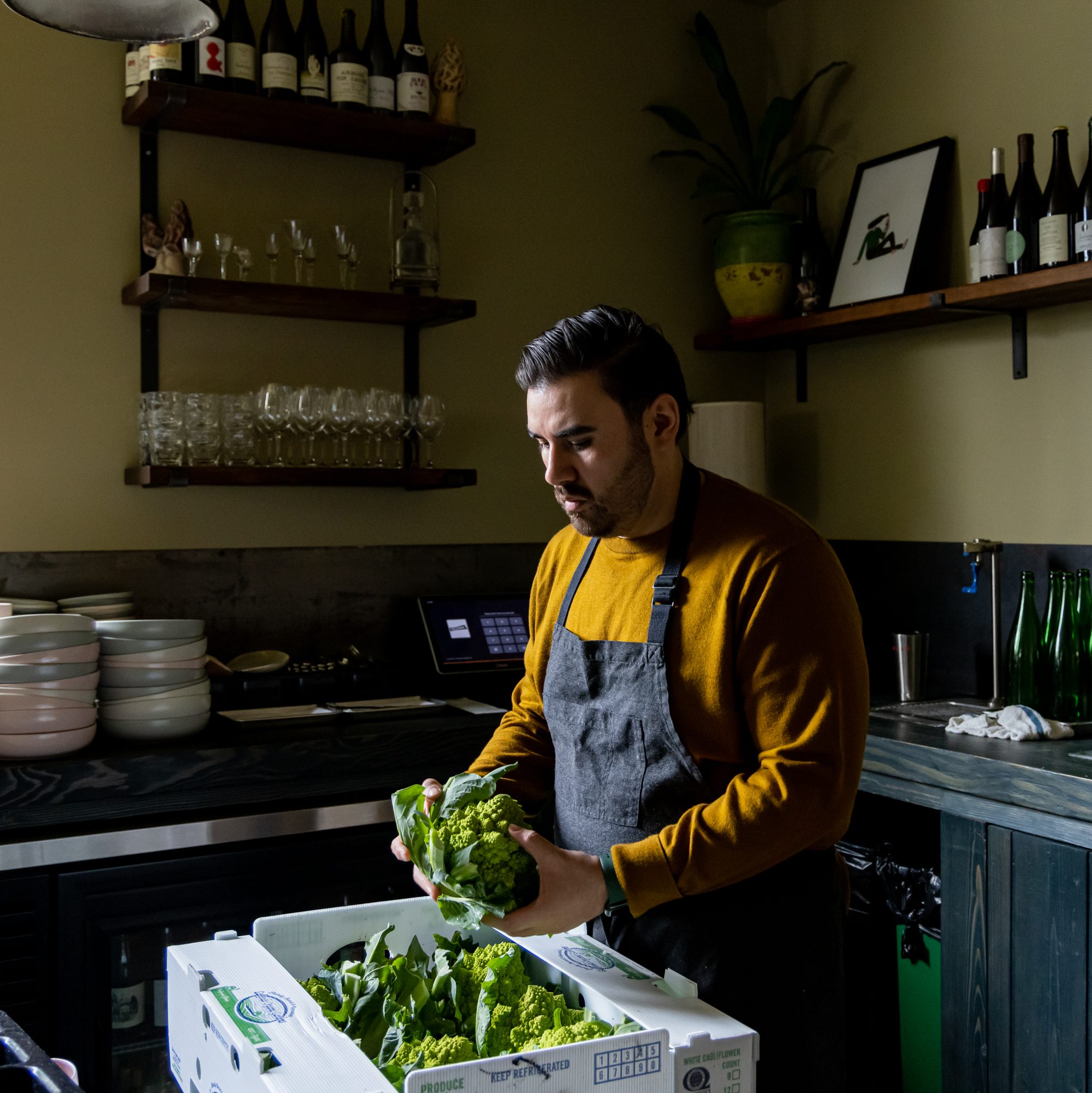 A man in a gray apron and mustard sweater stands in a kitchen holding fresh romanesco. Shelves with glasses and bottles are in the background.