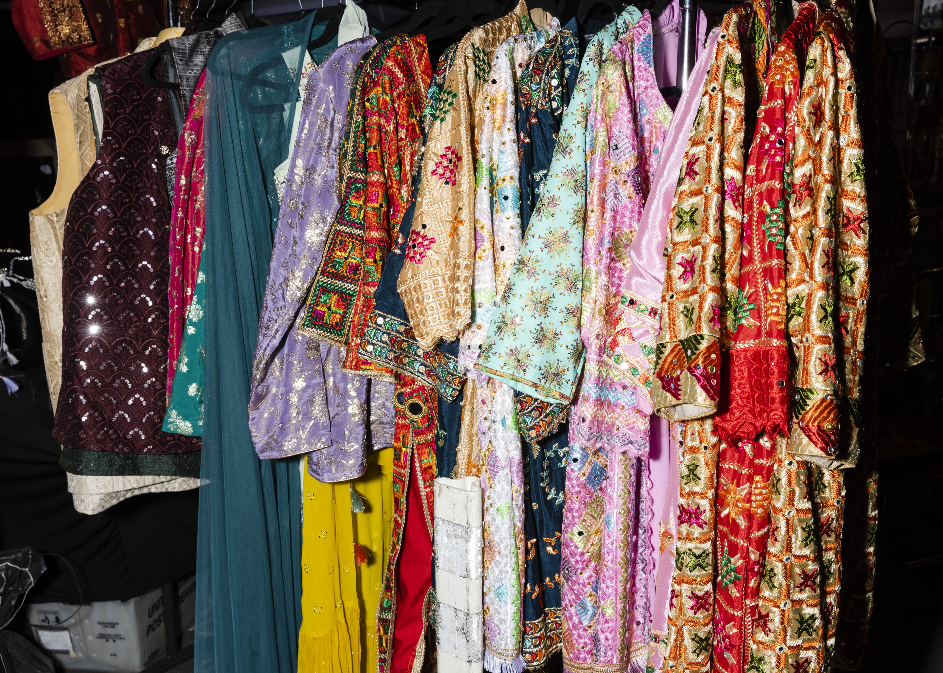 The image shows a row of colorful, intricately embroidered clothes hanging on a rack. The fabrics are vibrant, featuring reds, greens, blues, and gold designs.