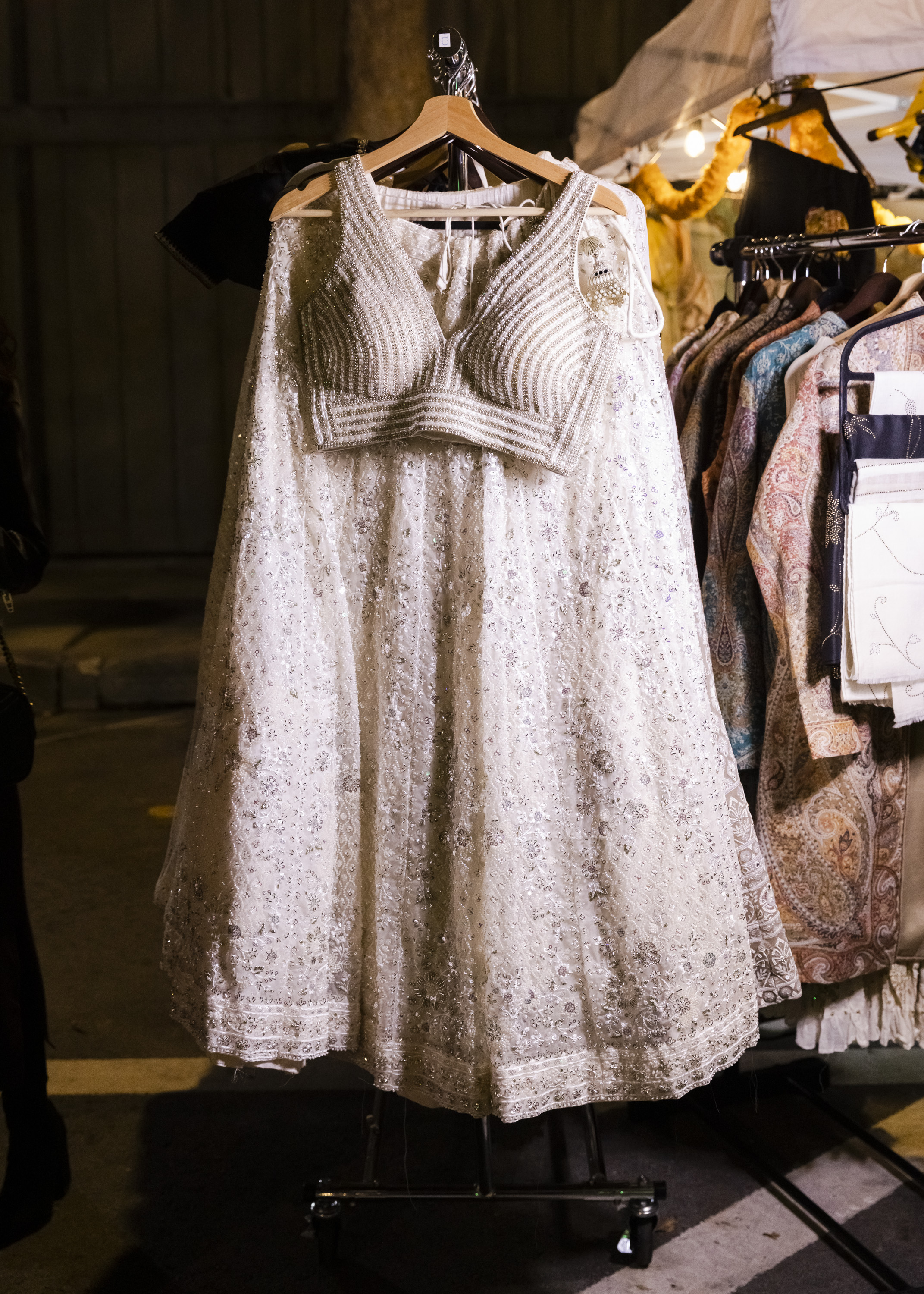 A sparkling, intricately embroidered white dress on a hanger is displayed at a market stall. It's surrounded by various colorful, patterned fabrics.