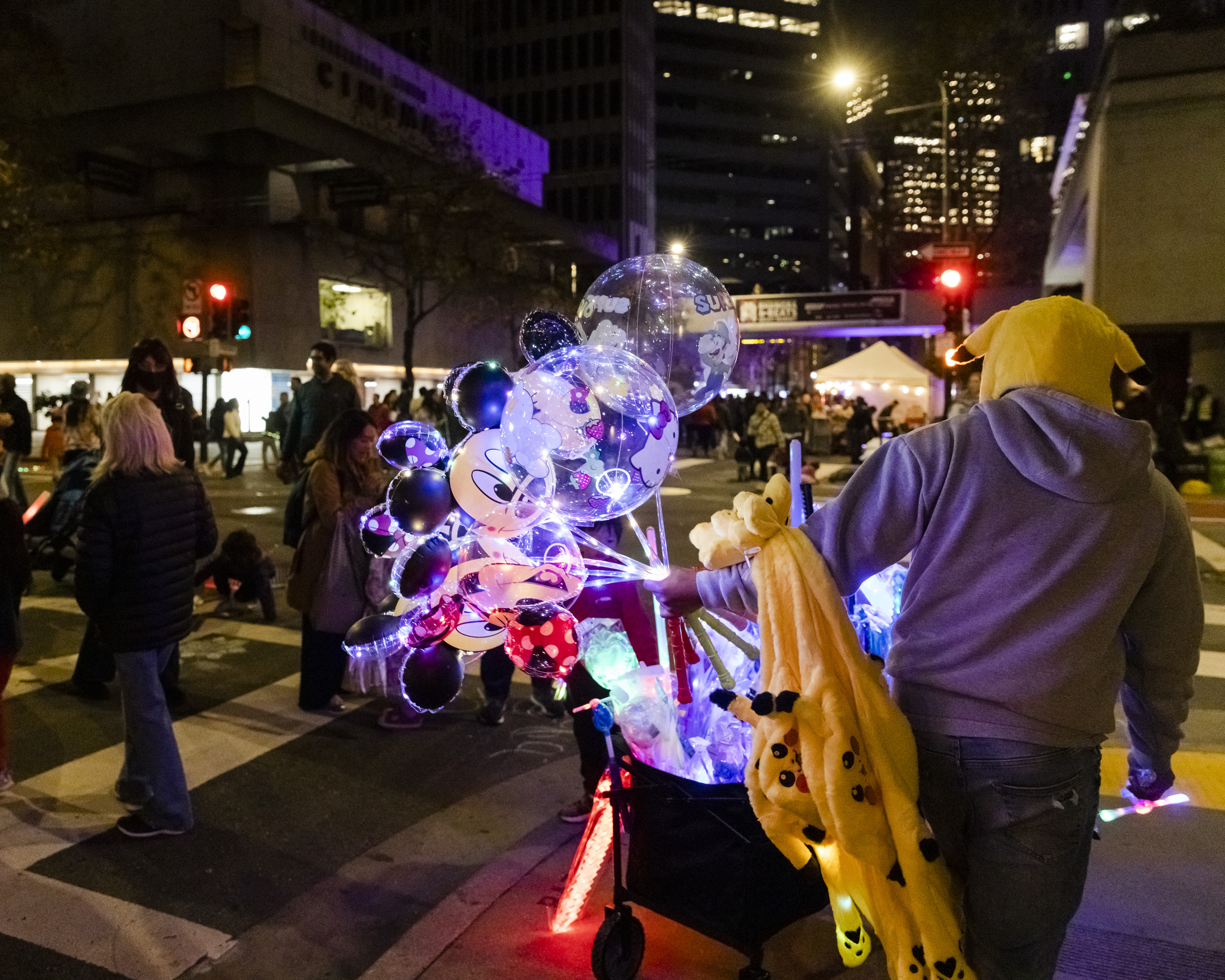 A street vendor at a night event sells glowing balloons and wears a Pikachu hoodie. People walk by under city lights, giving the scene a lively, festive atmosphere.