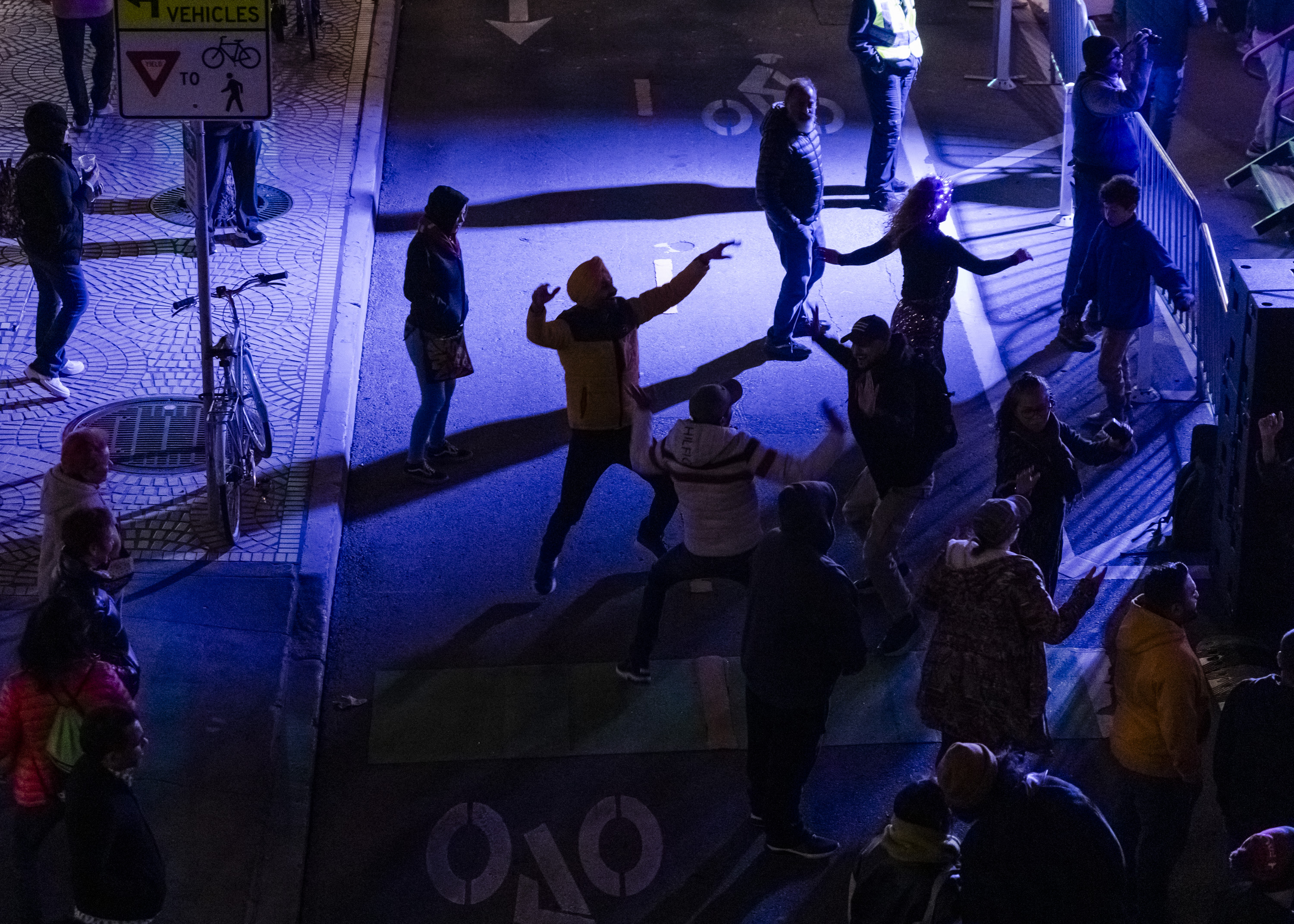 People are dancing on a street with blue and purple lights. A person in a yellow jacket stands out in the crowd. There are barriers and a bicycle nearby.