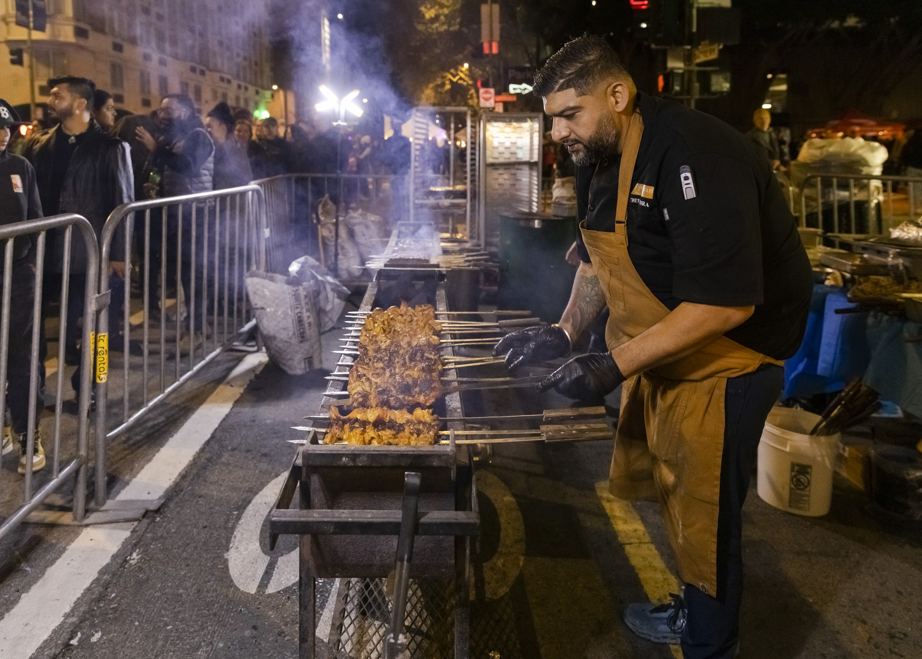 A man in an apron grills skewers of meat on a large outdoor barbecue at night, surrounded by smoky air and a curious crowd behind metal barriers.