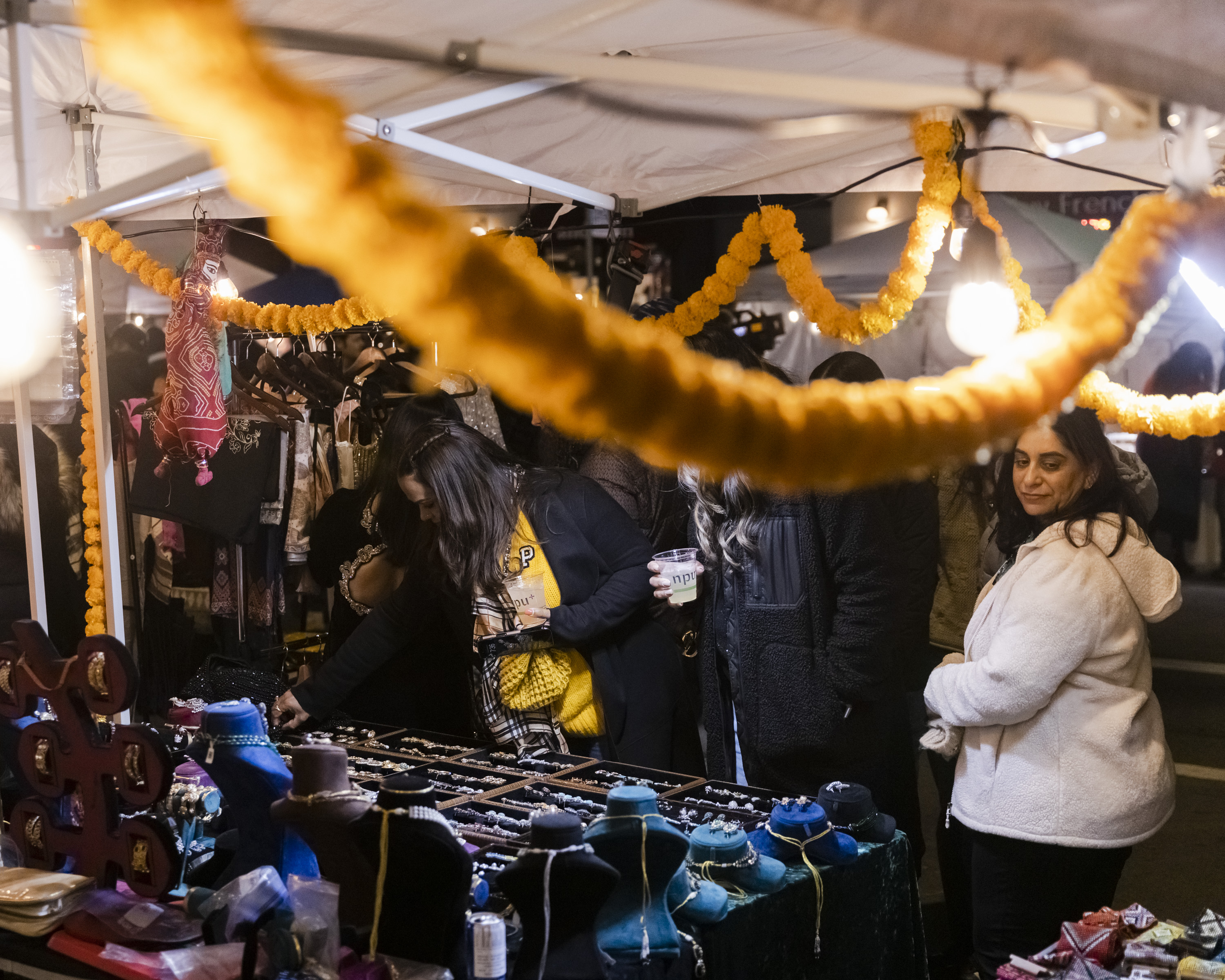 People are browsing a vibrant outdoor market stall at night, with garlands of marigold flowers hanging and various jewelry items displayed on tables.