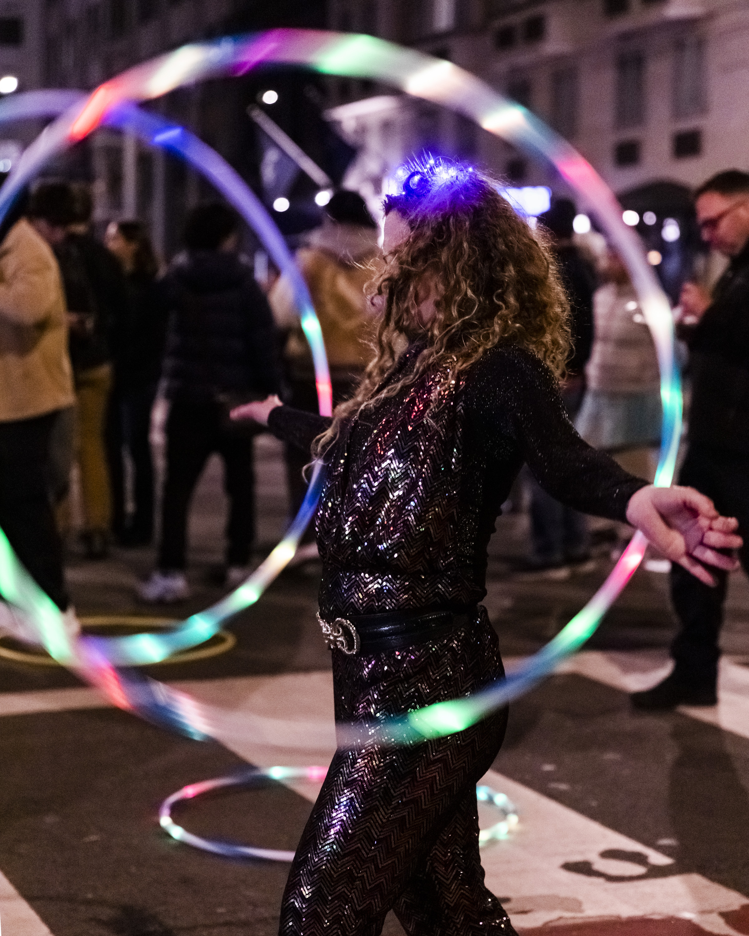 A person with curly hair spins a colorful LED hula hoop on a street at night, wearing a sparkly black outfit, surrounded by a blurred crowd.