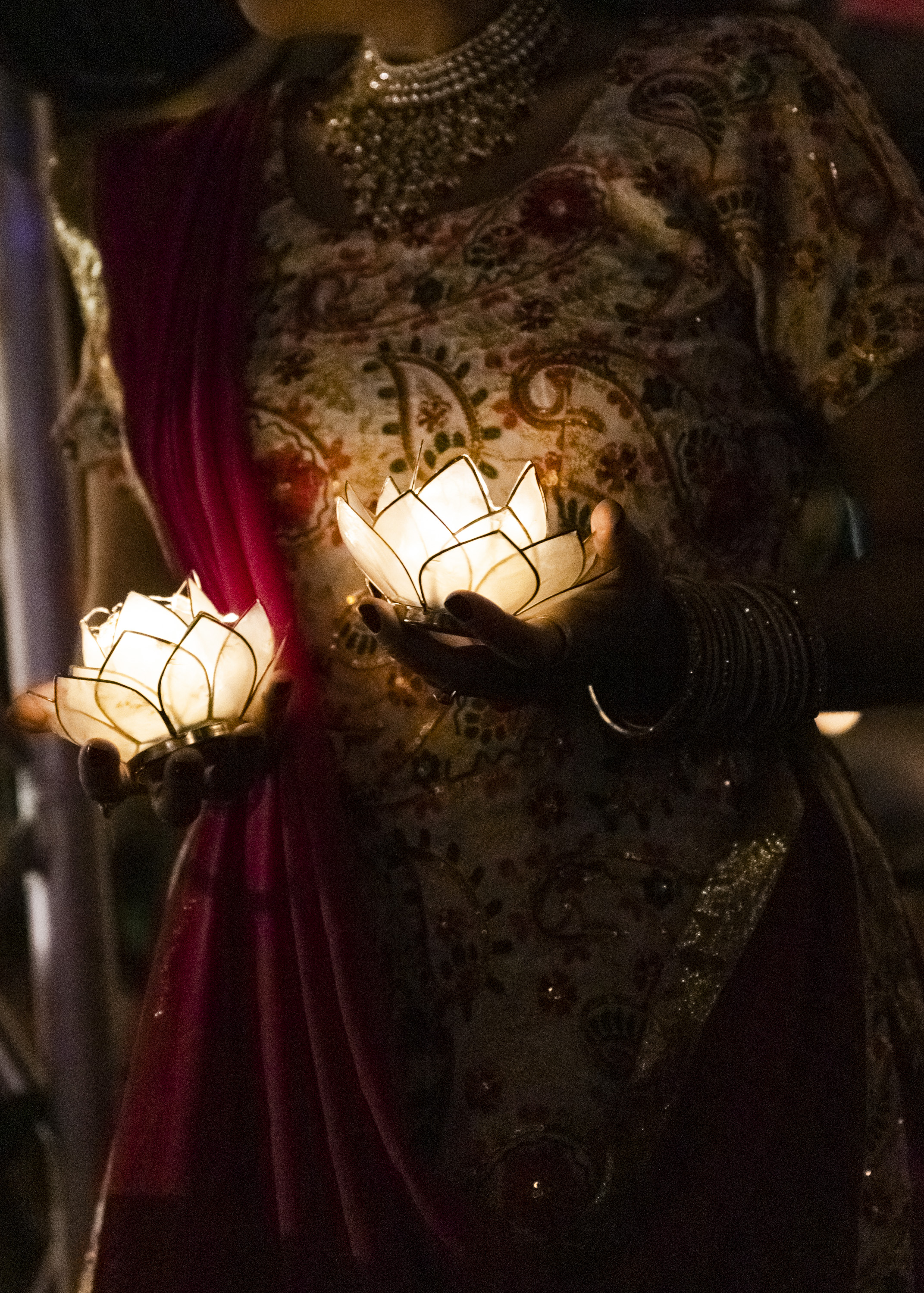 A person in traditional attire holds glowing lotus-shaped candles. The outfit is richly patterned with a maroon drape and sparkling jewelry.