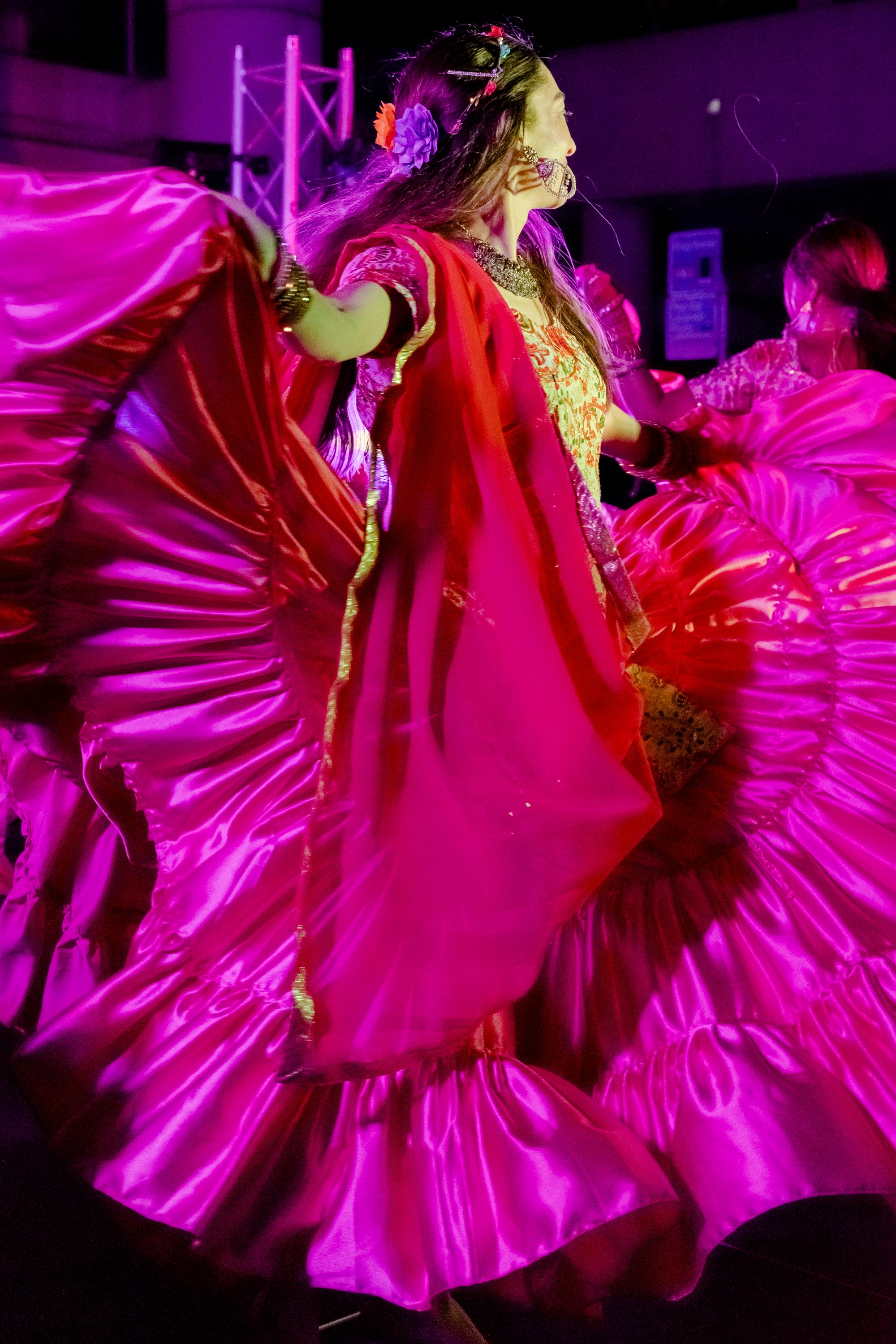 A dancer in vibrant traditional attire twirls, showcasing a wide, flowing magenta skirt under bright stage lights, creating a dynamic and colorful scene.