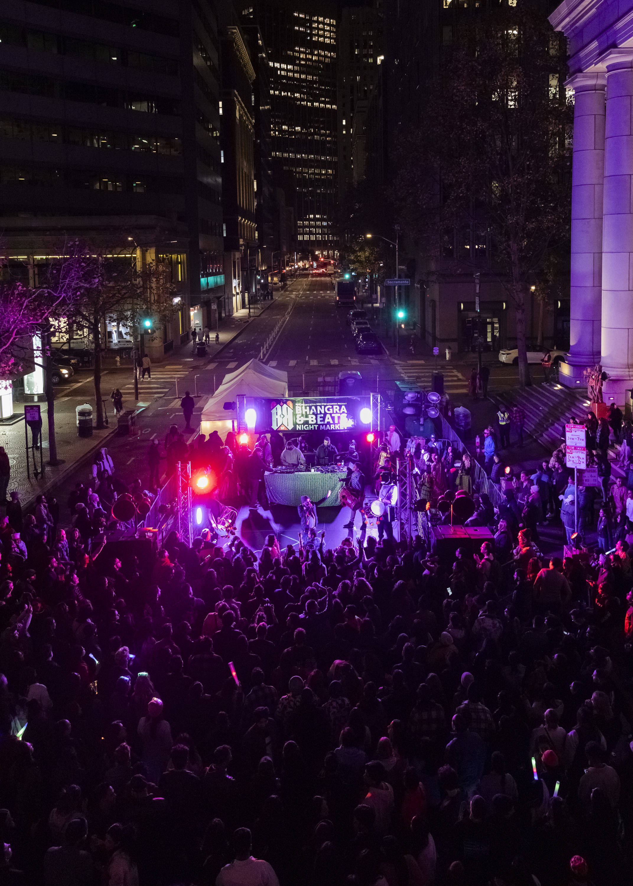 A vibrant night scene shows a crowded street market event with performers on stage, colorful lights, and a backdrop of city buildings at night.