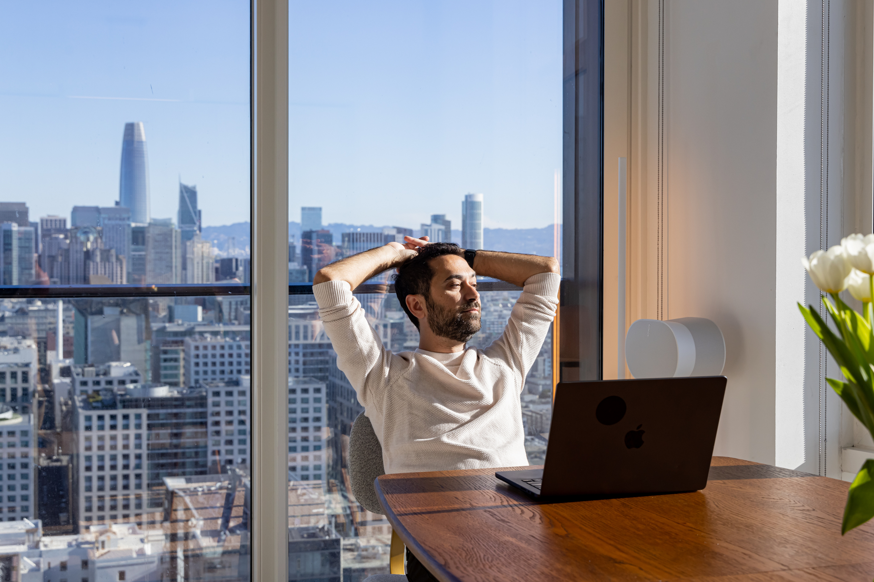 A man relaxes in an office with a laptop on a wooden table, overlooking a city skyline through large windows. Tulips are on the table, and sunlight streams in.