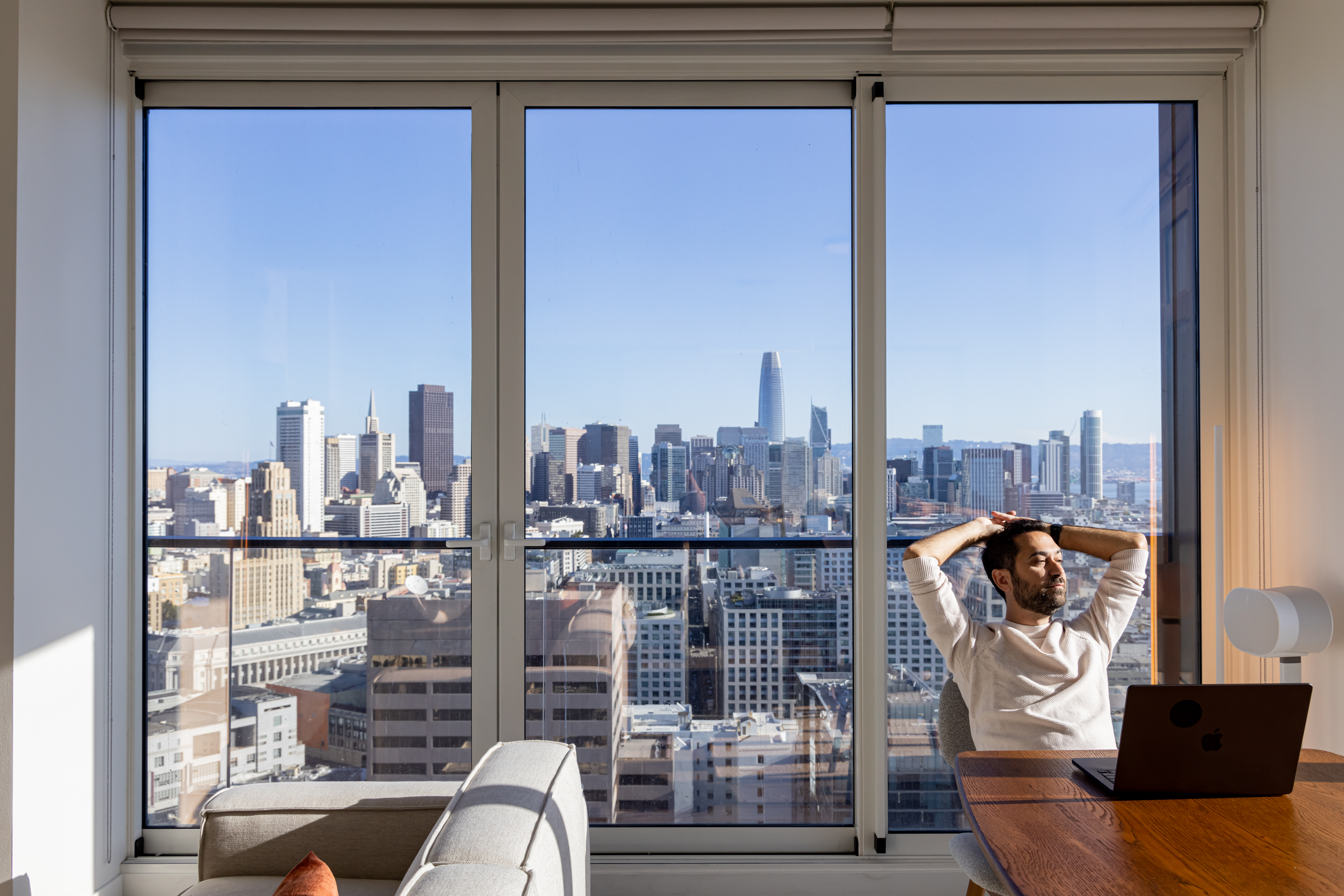 A man sits at a table with a laptop, looking relaxed, in front of a large window showcasing a clear city skyline with tall buildings.