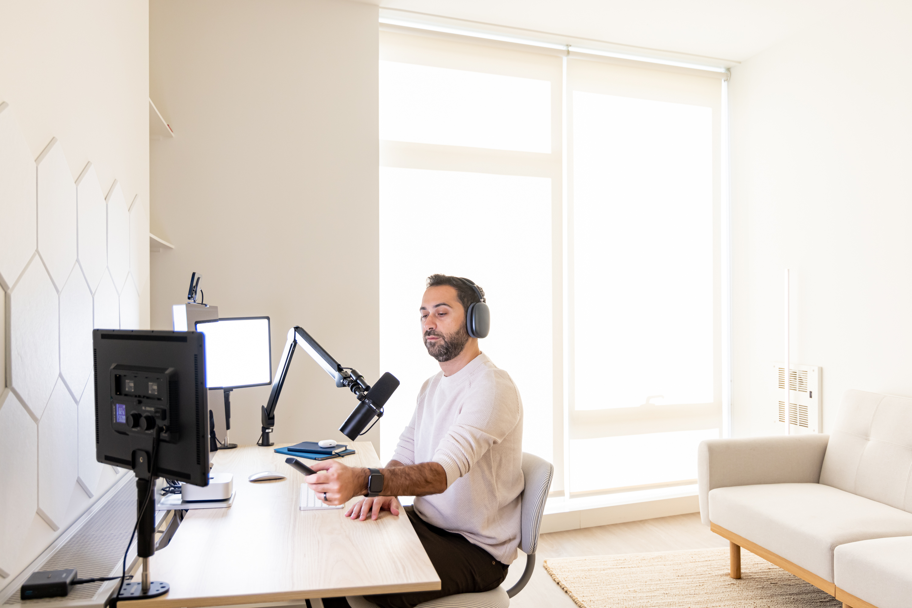 A person sits at a desk in a bright room, wearing headphones, using a computer and microphone, with a modern sofa and large window in the background.