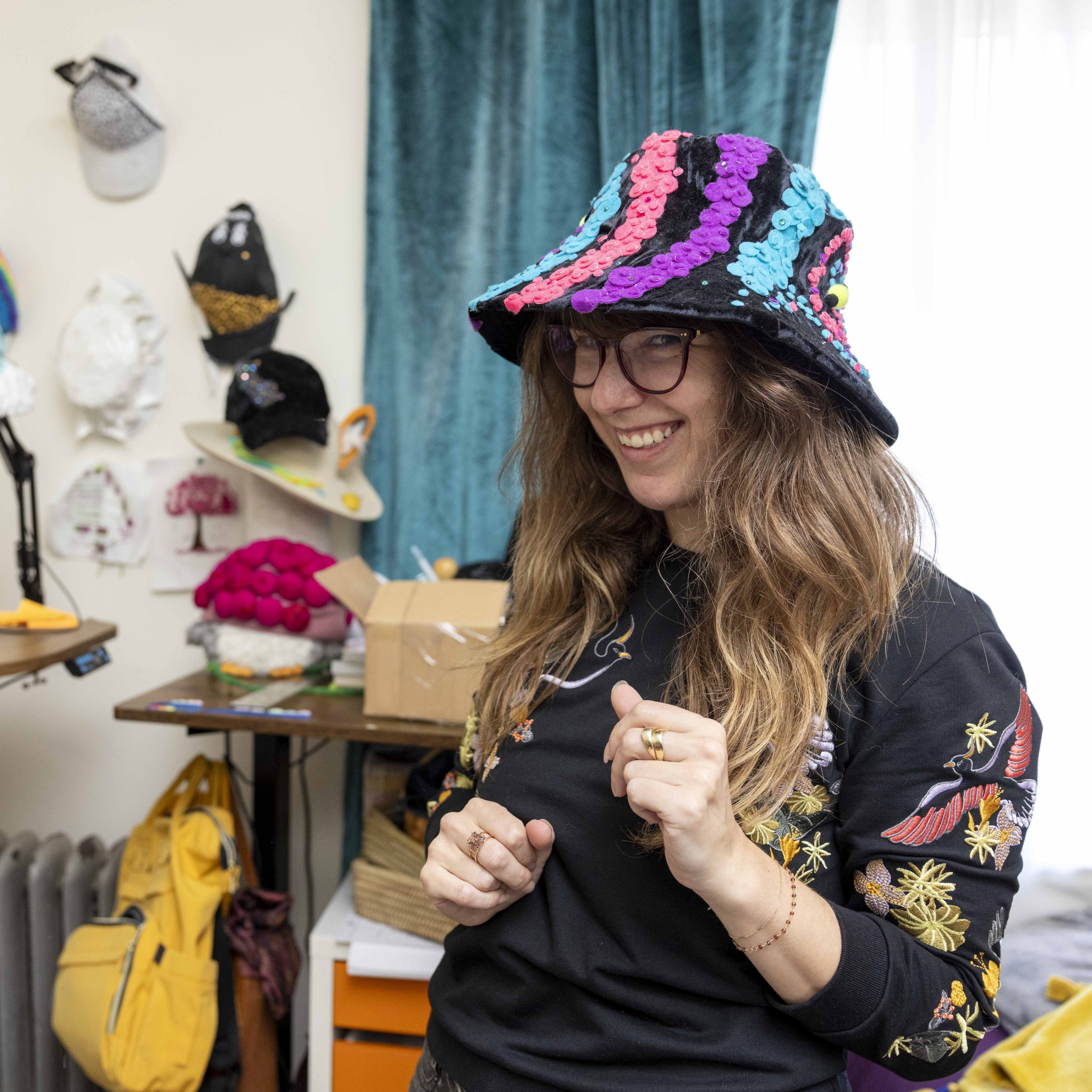 A person smiles while wearing a colorful, sequined hat in a room filled with various hats and craft items. They have long hair and are wearing glasses and a patterned shirt.