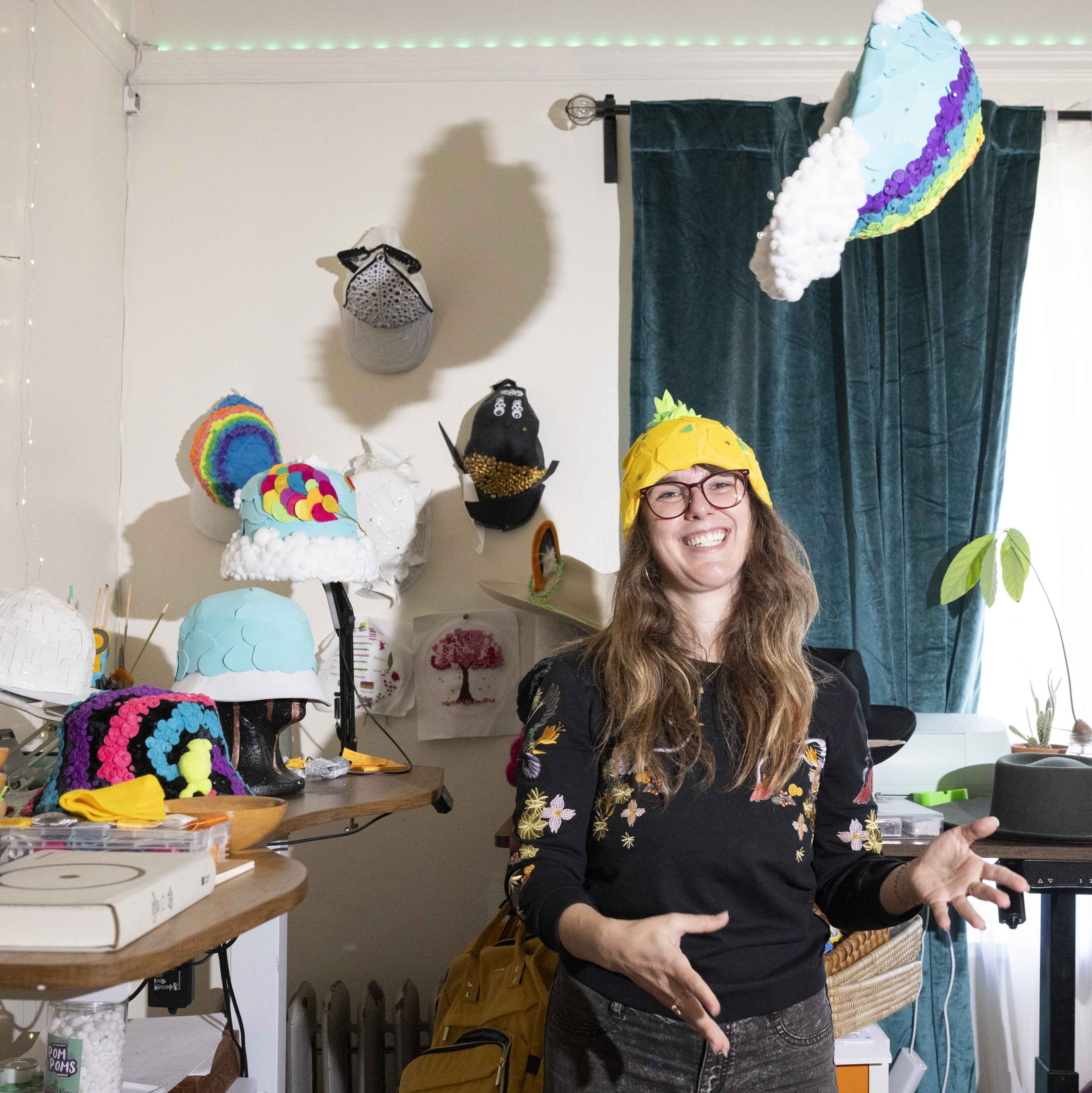 A smiling person with long hair stands in a colorful room. They're wearing a unique hat, surrounded by various creative hats on a table and the wall.